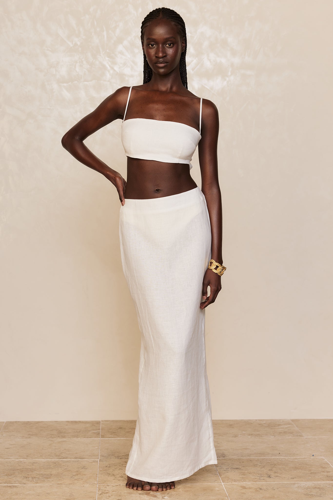 Model wearing white bandeau top and long white skirt, barefoot, braided hair, gold chain-link bracelet on right wrist.
