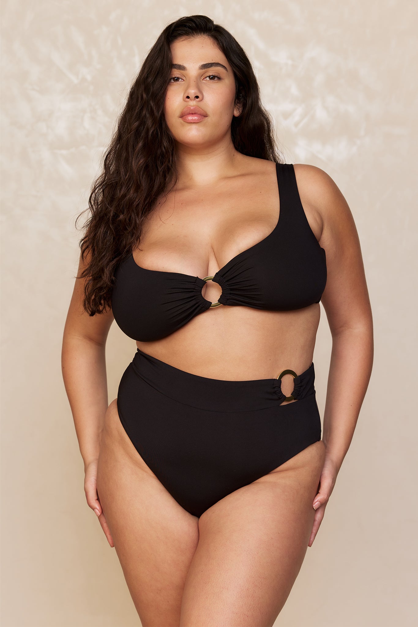 Plus-size model wearing black two-piece swimsuit with gold circular rings at center and hip, neutral studio backdrop.