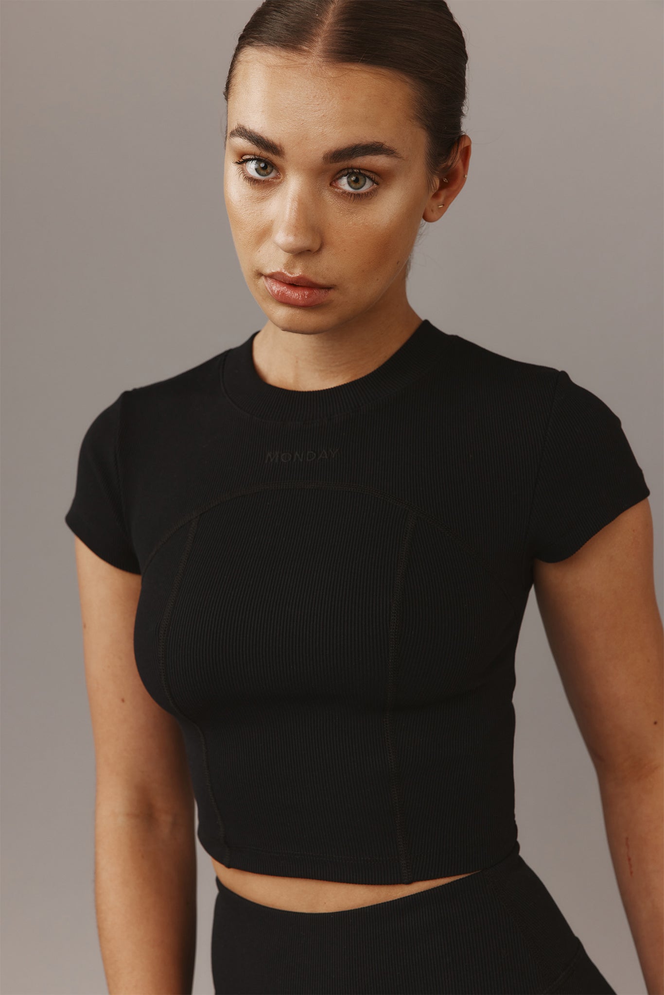 Head-and-shoulders portrait of a young woman wearing a black ribbed short-sleeve crop top stamped 'MONDAY' on the chest.