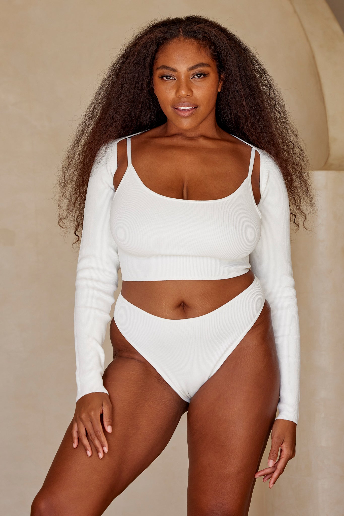 Model wearing white ribbed long-sleeve crop top with thin straps and matching high-cut briefs, standing against a neutral backdrop.