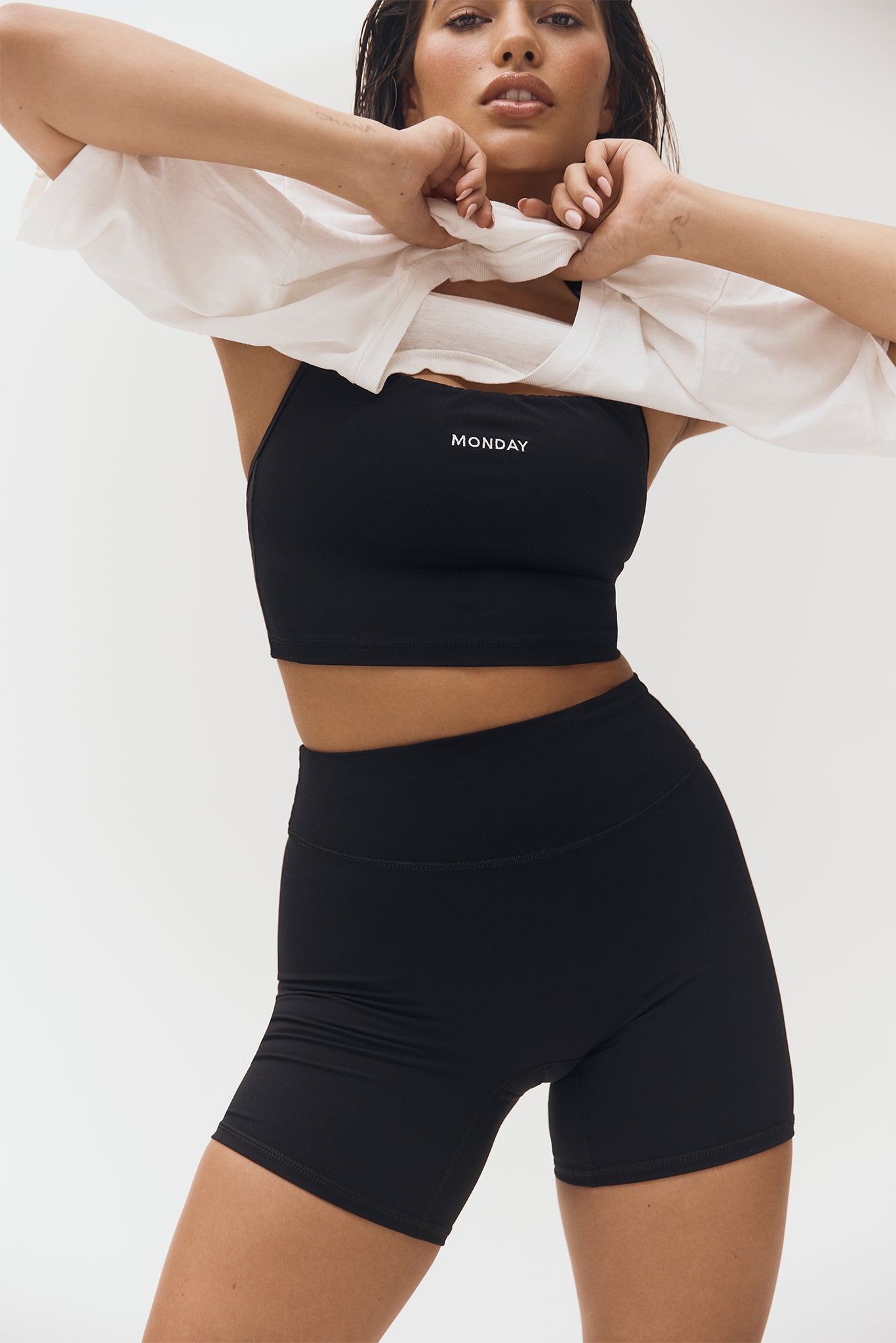 Woman wearing black crop top with MONDAY printed on the chest and high-waisted black bike shorts, lifting a white shirt
