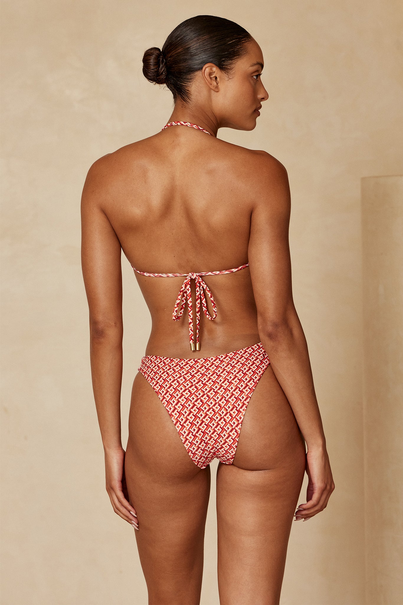 Back view of model wearing red geometric high-leg bikini bottoms and tied halter top, hair in a low bun, beige backdrop.