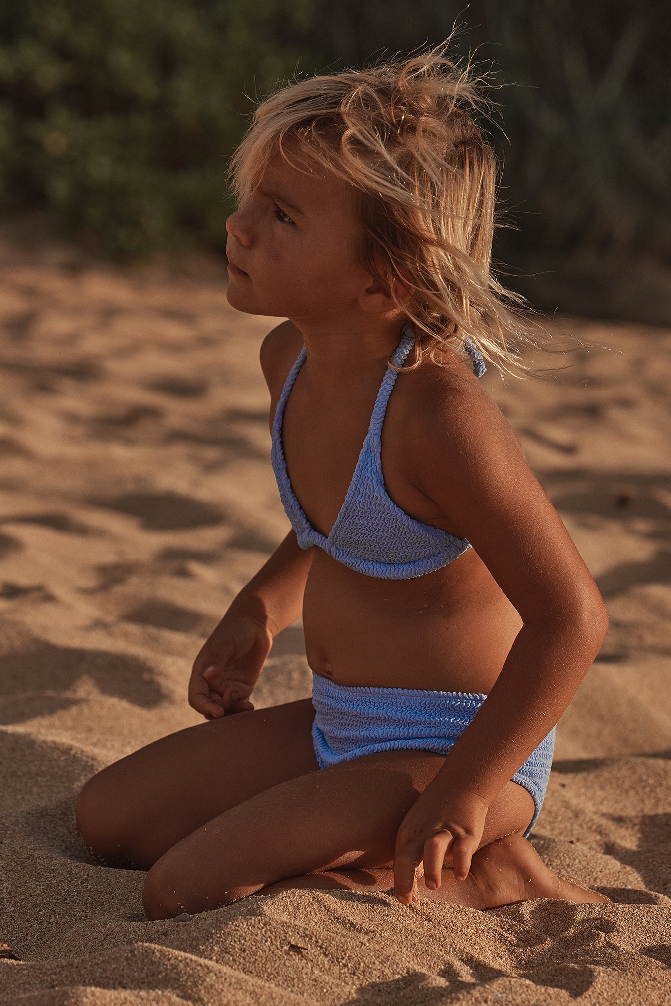 Child kneeling on sandy beach wearing a light blue knitted bikini, windblown hair, looking up to the left.