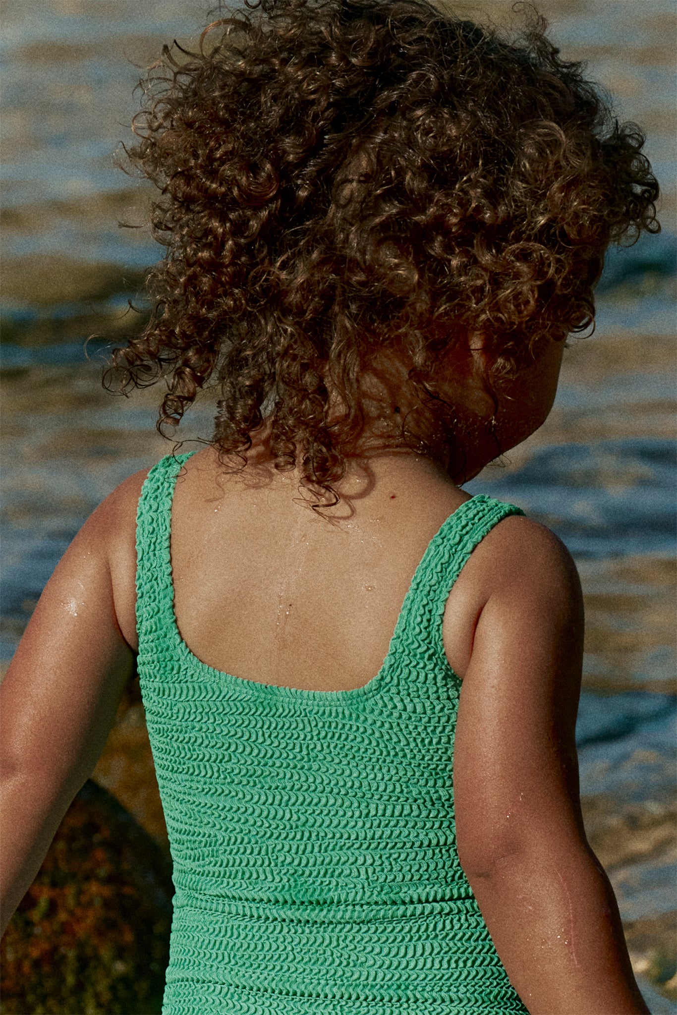 Back view of a young child with curly hair wearing a green textured swimsuit standing at the water's edge