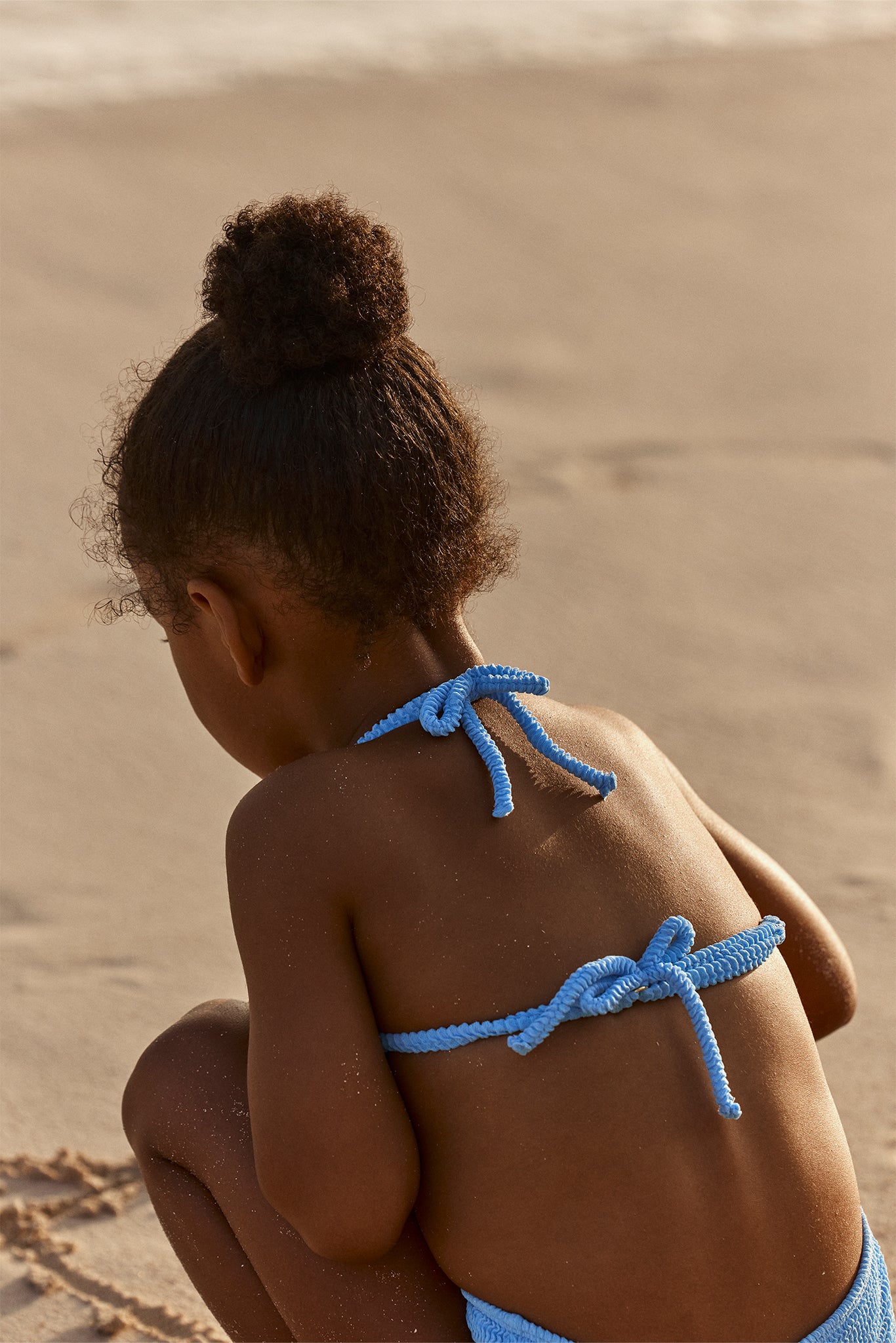 Young child sitting on sand with back turned, hair in a bun, wearing a blue bikini with rope-tie straps.