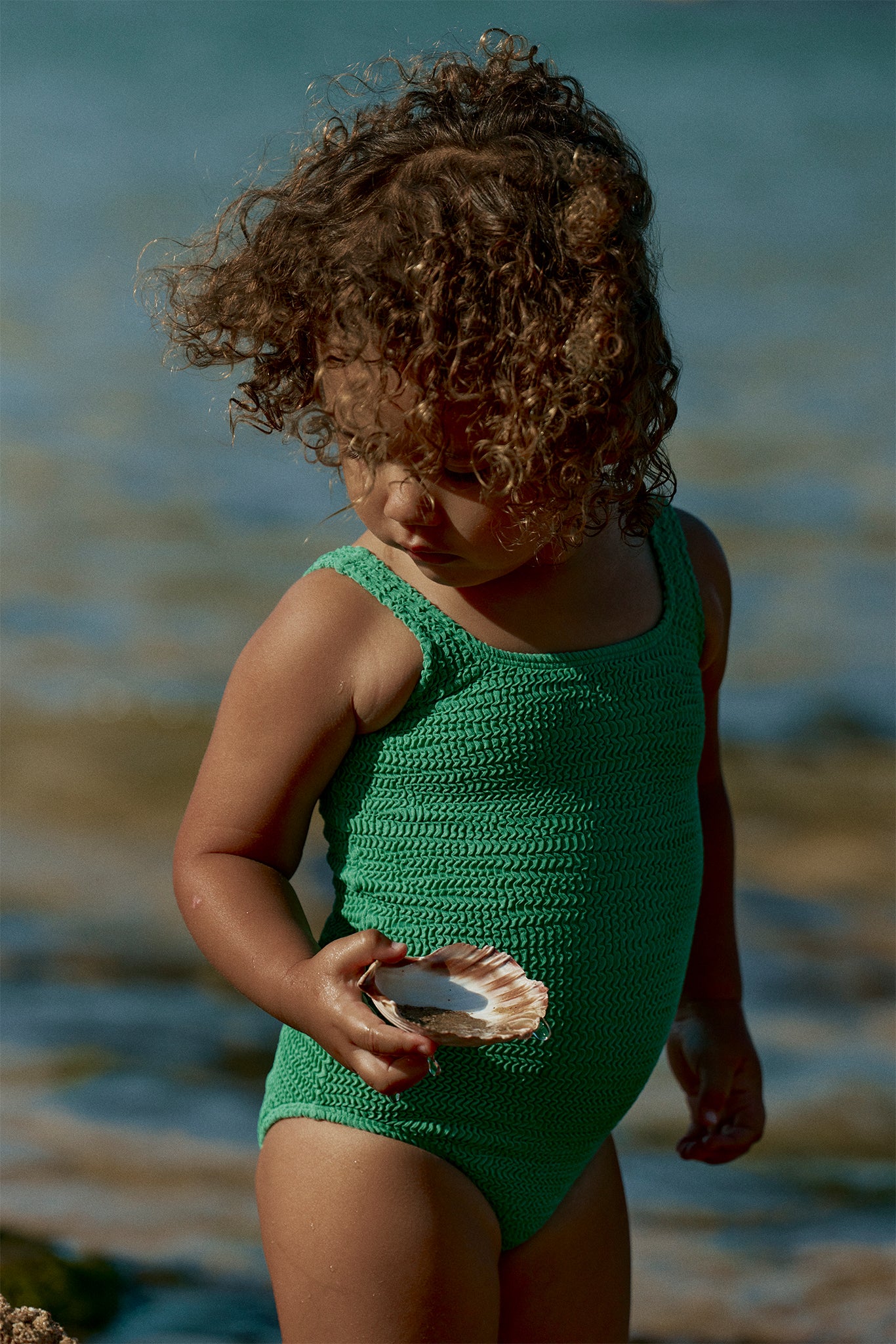 Child in a green knit swimsuit holding a scallop shell at the shoreline, looking down.