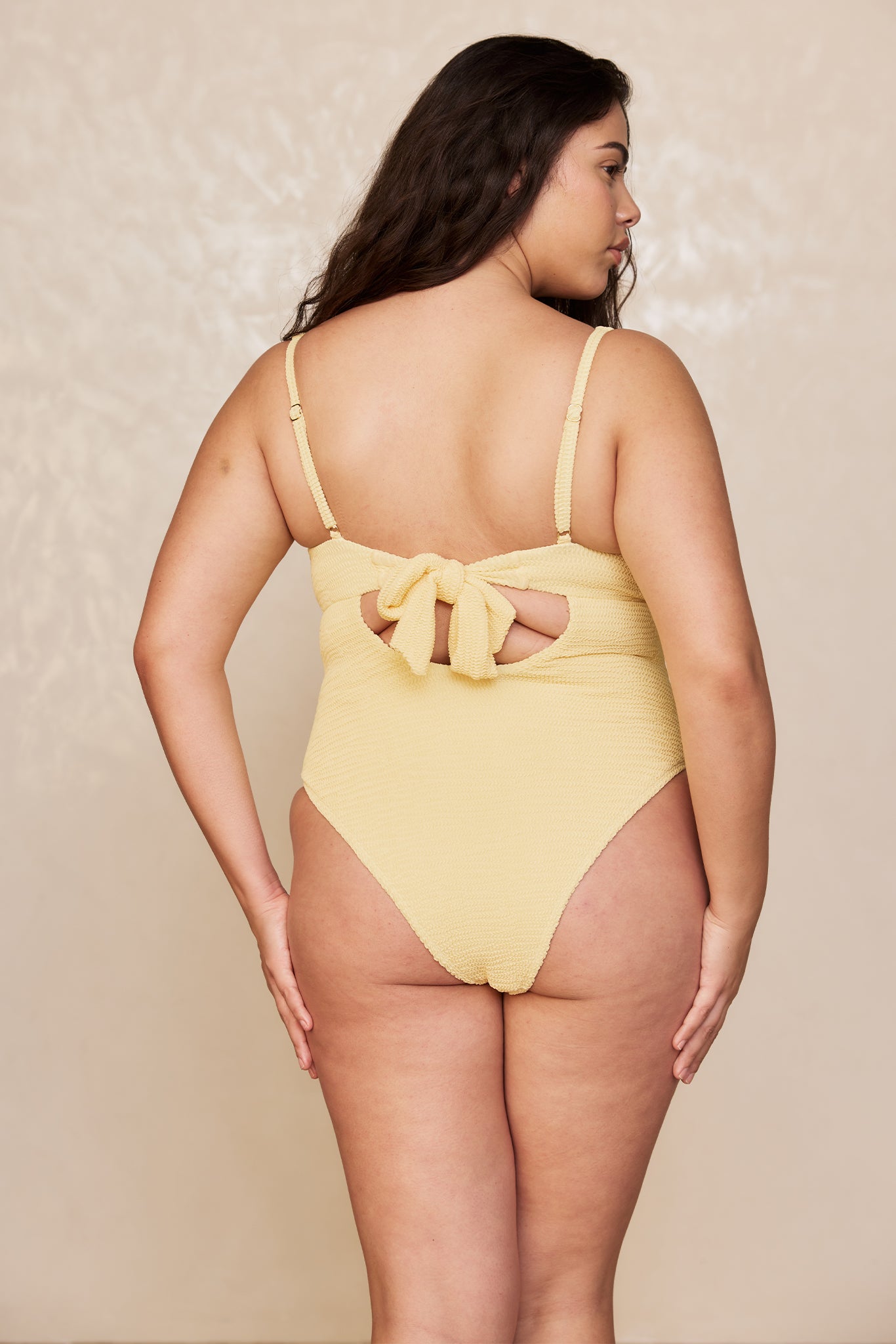 Back view of plus-size model wearing textured pale yellow one-piece swimsuit with tied bow at back.