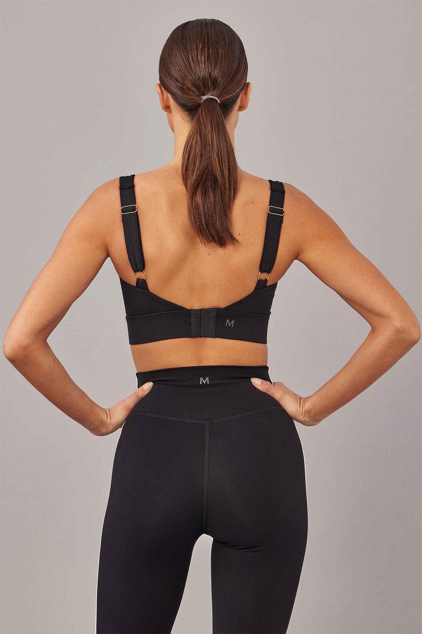 Back view of woman wearing black sports bra and high-waist leggings with visible M logos, low ponytail, hands on hips.