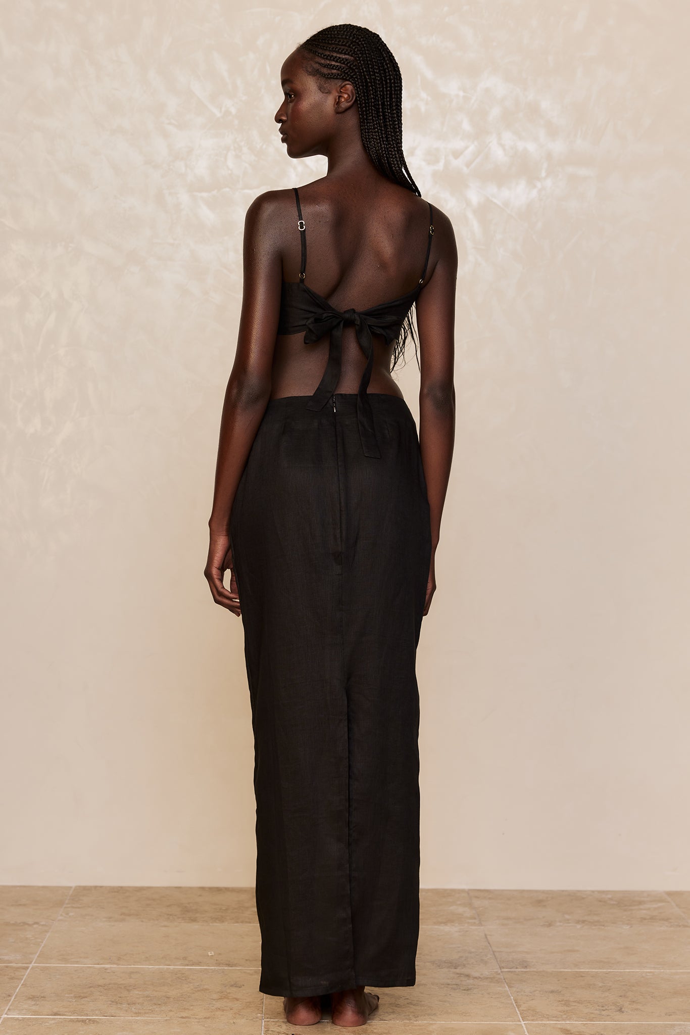 Rear view of a model with braided hair wearing a black tie-back crop top and long black maxi skirt, standing barefoot.