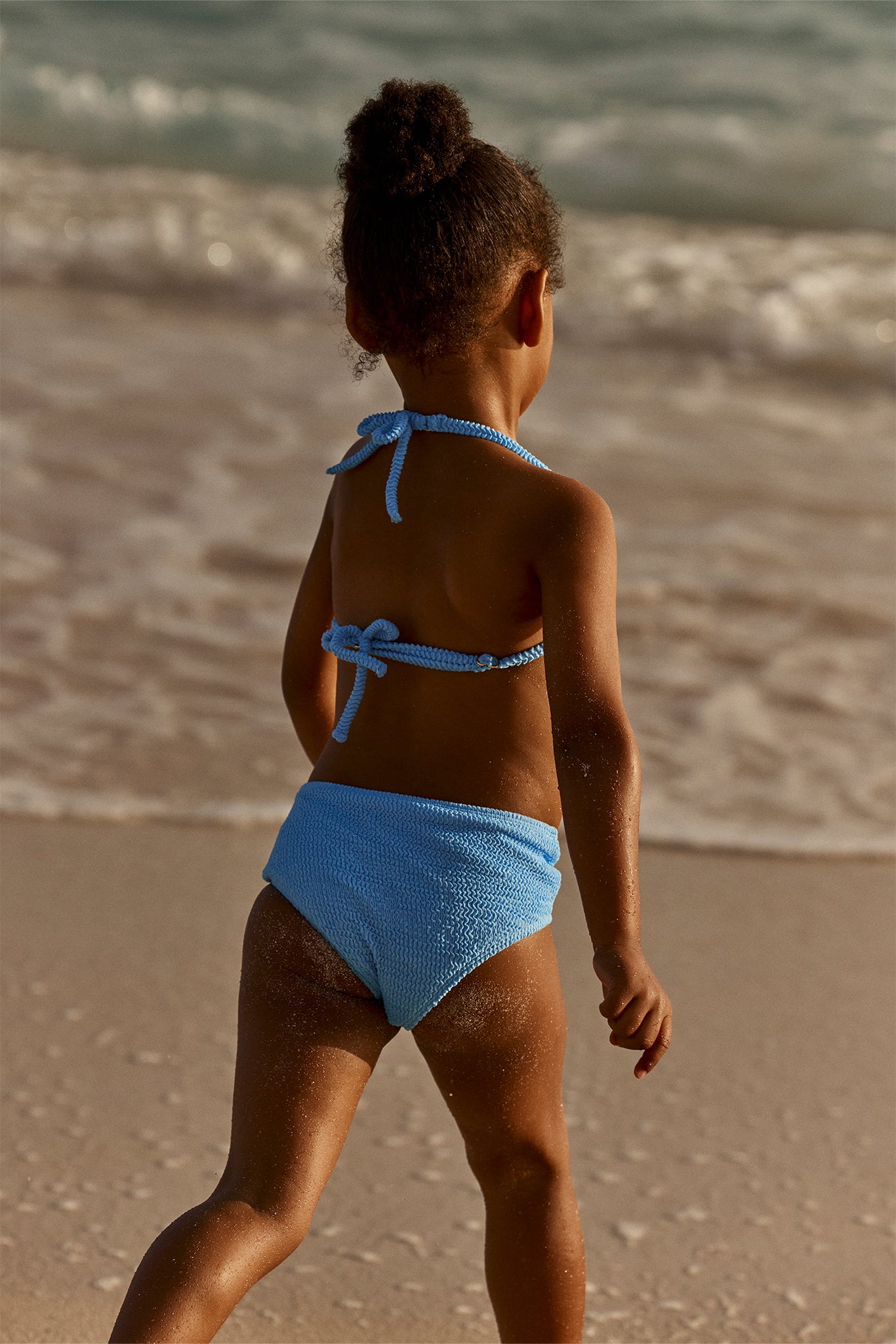 Young child on sandy beach facing waves, wearing light-blue textured two-piece swimsuit.