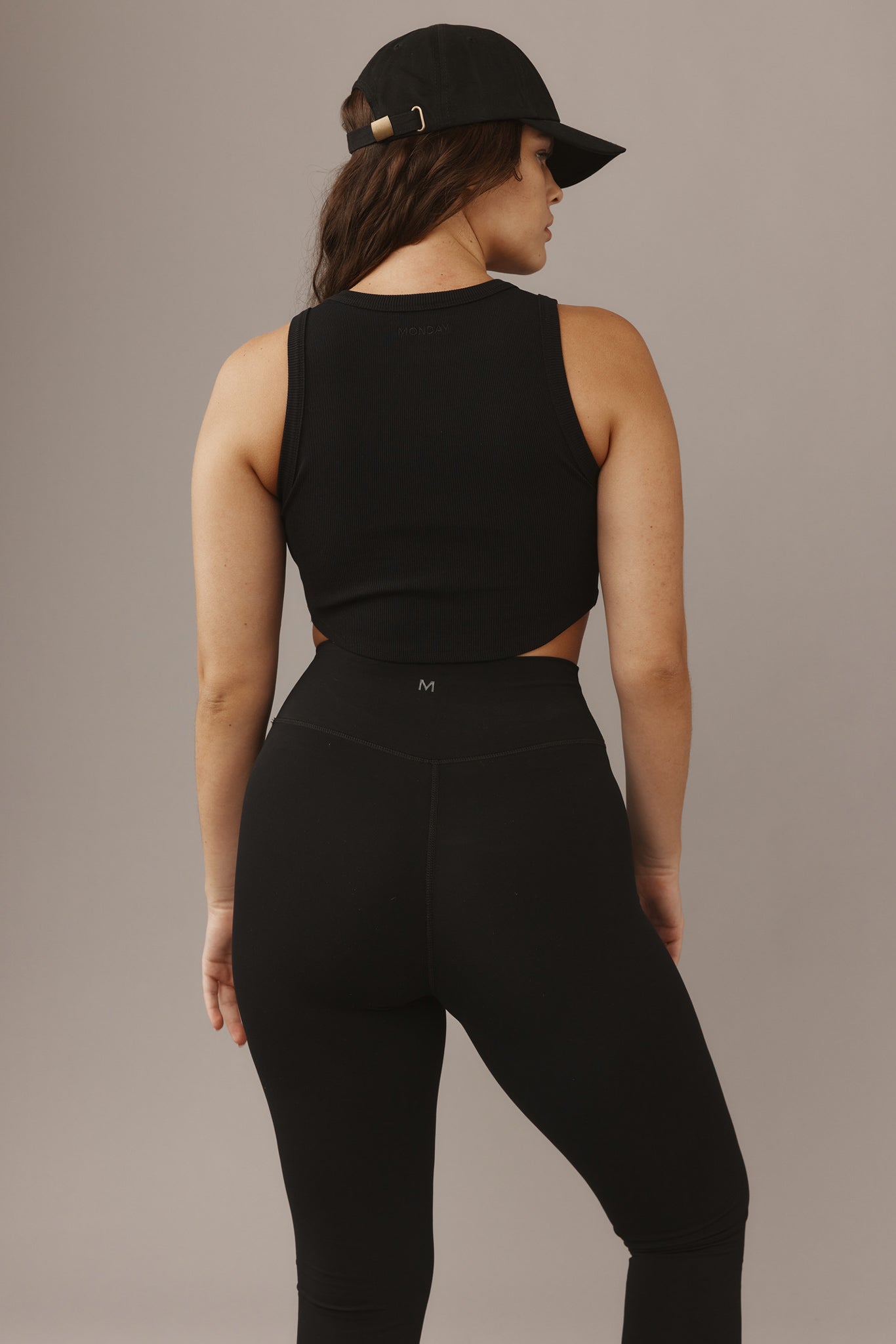Back view of woman wearing black ribbed tank ('MONDAY' at neckline), high-waist black leggings with 'M' logo, and black cap