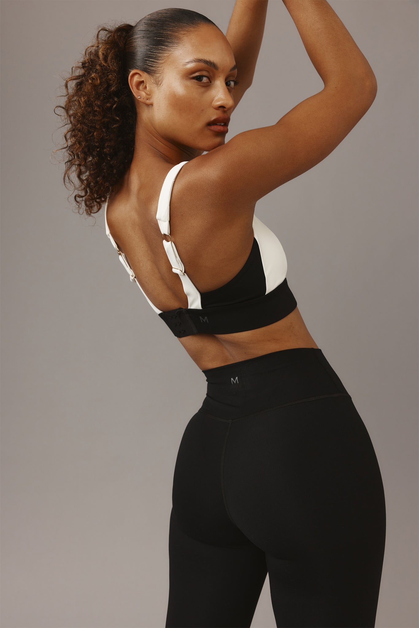 Back view of a woman raising her arms wearing a white sports bra and high-waist black leggings, 'M' printed on waistband.