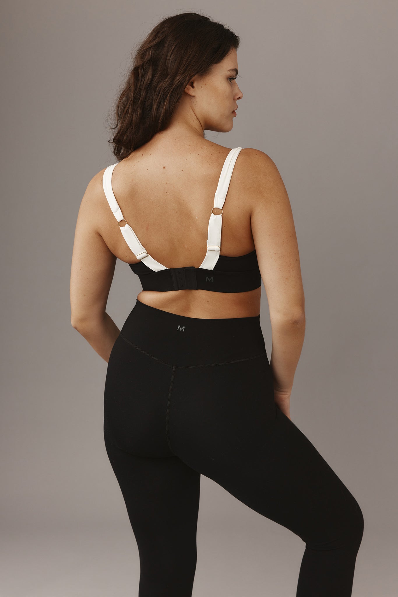 Rear view of a woman wearing a black sports bra with white adjustable straps and high-waist black leggings with M logo.