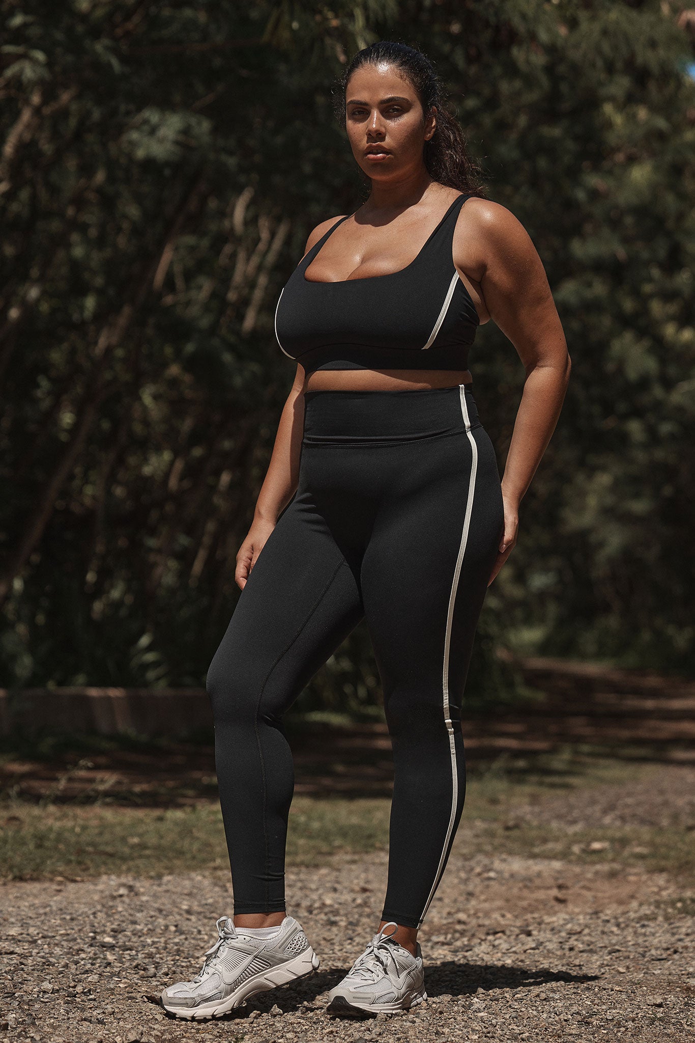 Woman standing on a gravel path wearing black sports bra, black leggings with white side stripes, and white sneakers.