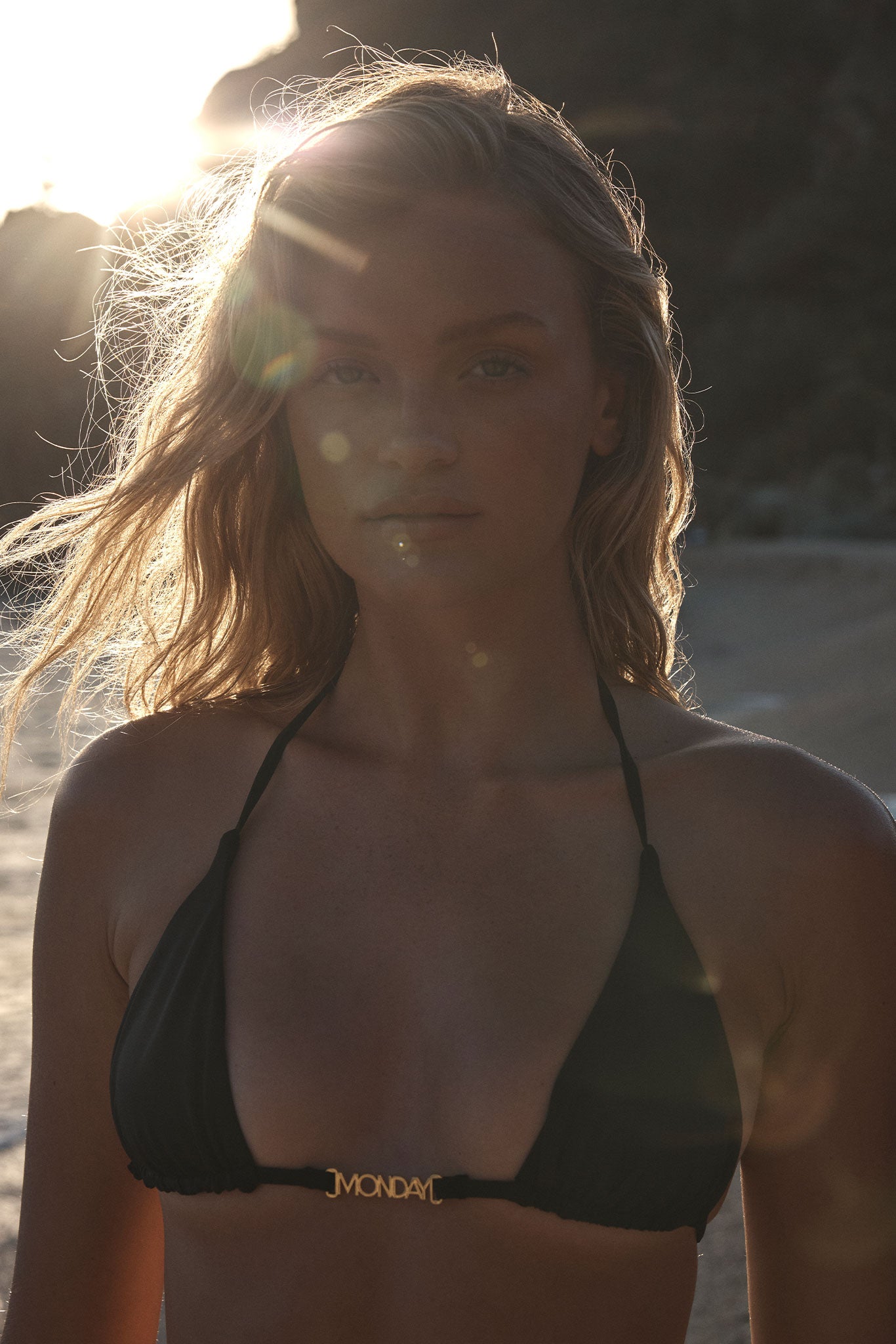 Young woman backlit on a beach wearing a black triangle bikini top with gold clasp reading 'MONDAY'; sun flare across face.