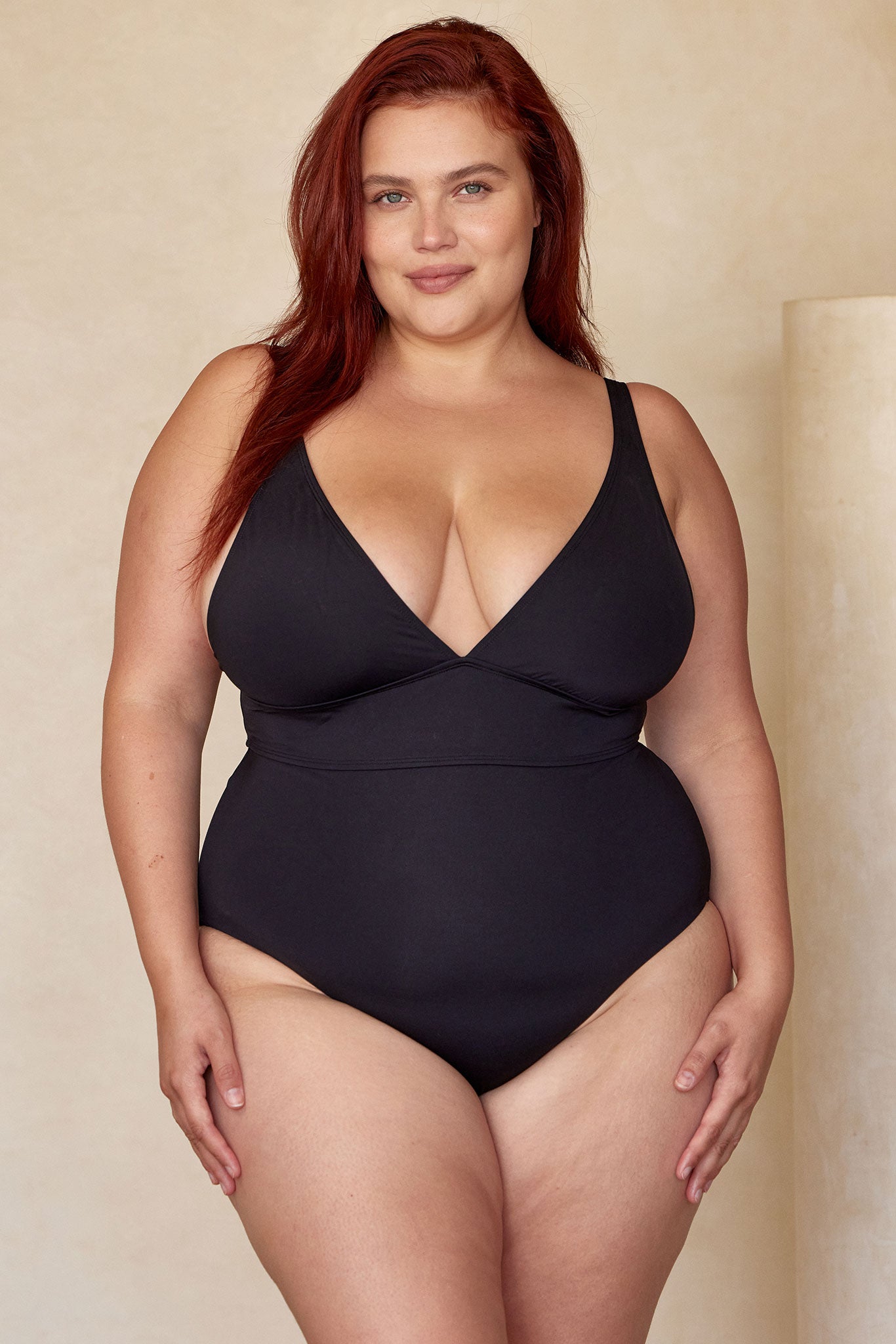 Plus-size woman with red hair wearing a black V-neck one-piece swimsuit, standing and smiling at the camera.