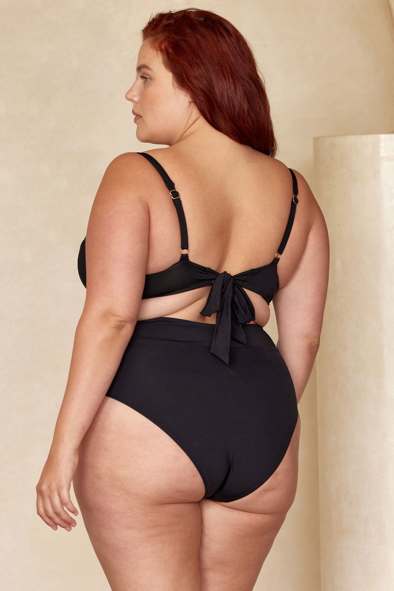 Plus-size woman shown from back wearing black tie-back bikini top and high-waist bikini bottoms.