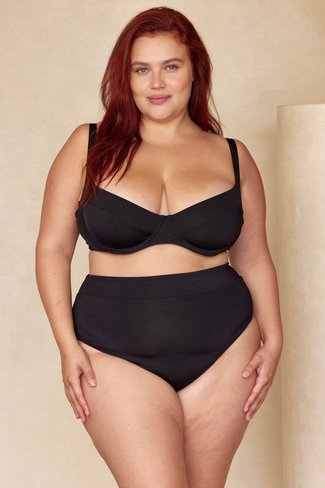 Plus-size red-haired model in black underwire bra and high-waist briefs standing against a beige backdrop, hands at sides.