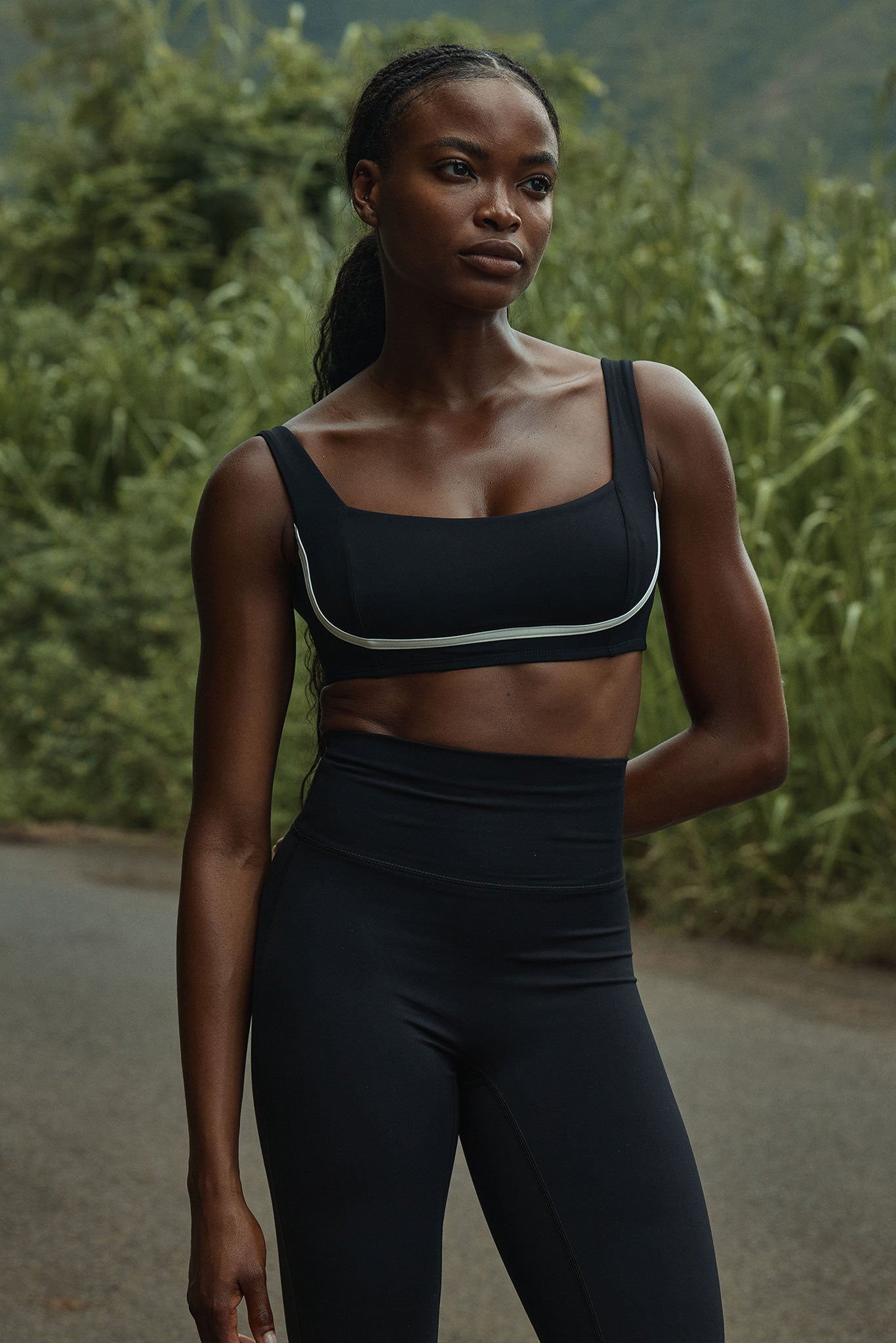 Woman in black sports bra and high-waist leggings standing on a road with tall green grass.