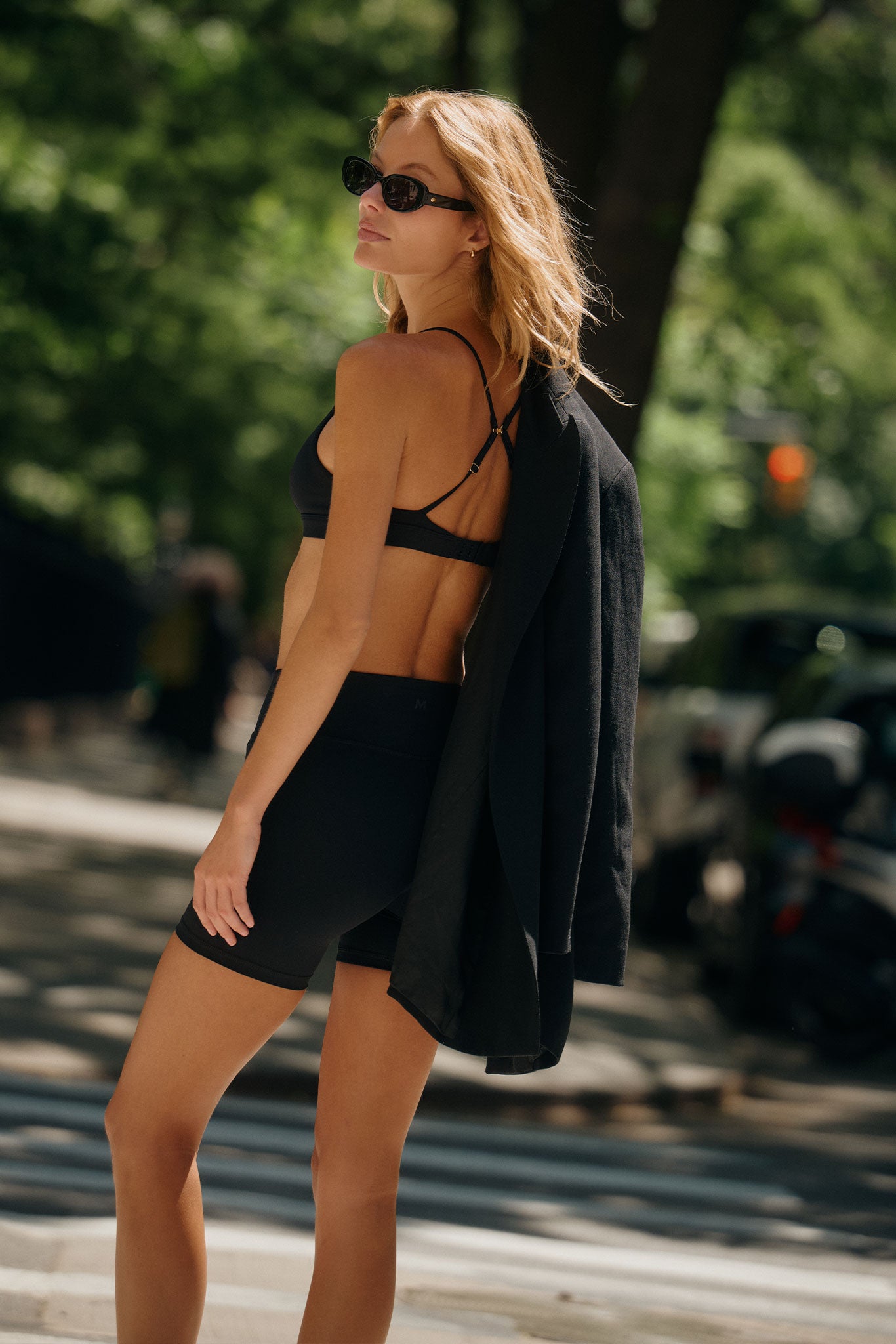 Woman in sunglasses wearing black sports bra and bike shorts with blazer draped over her shoulder on a sunlit city sidewalk