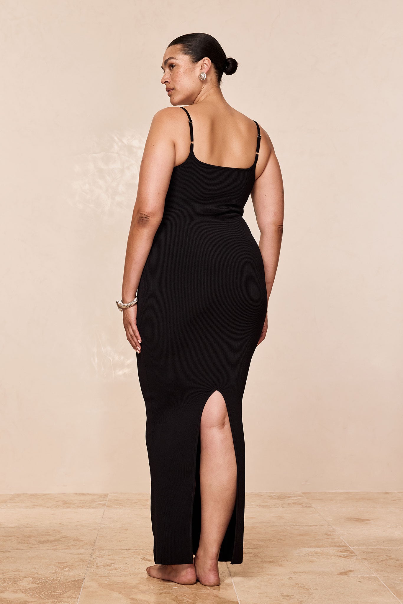 Model shown from behind wearing a black ribbed maxi dress with thin straps and a calf‑length back slit