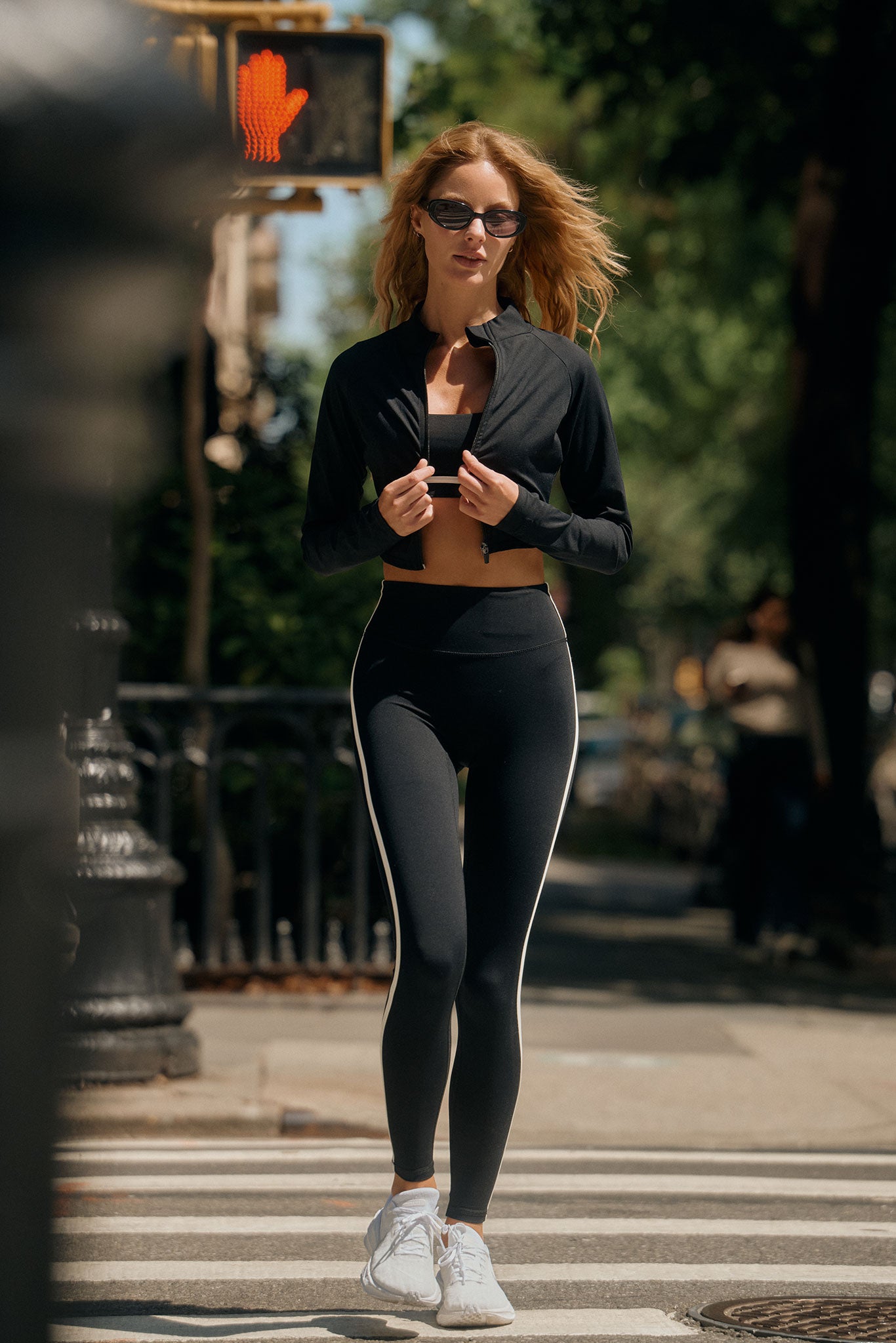 Woman in black athletic jacket and leggings walking across a city crosswalk; red raised-hand pedestrian signal visible overhead
