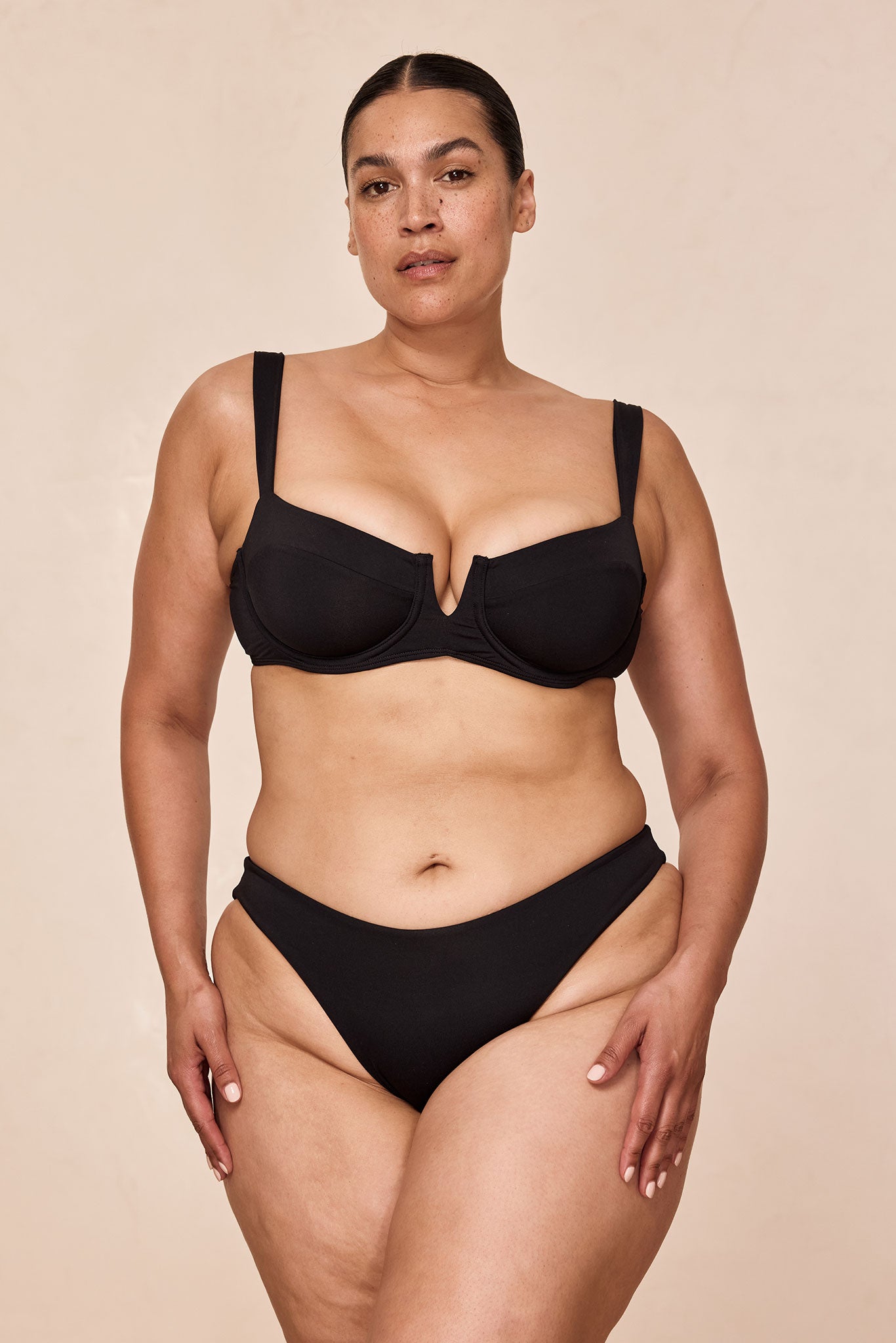 Plus-size model standing in black underwire bra and high-cut black bikini bottoms against a beige backdrop.