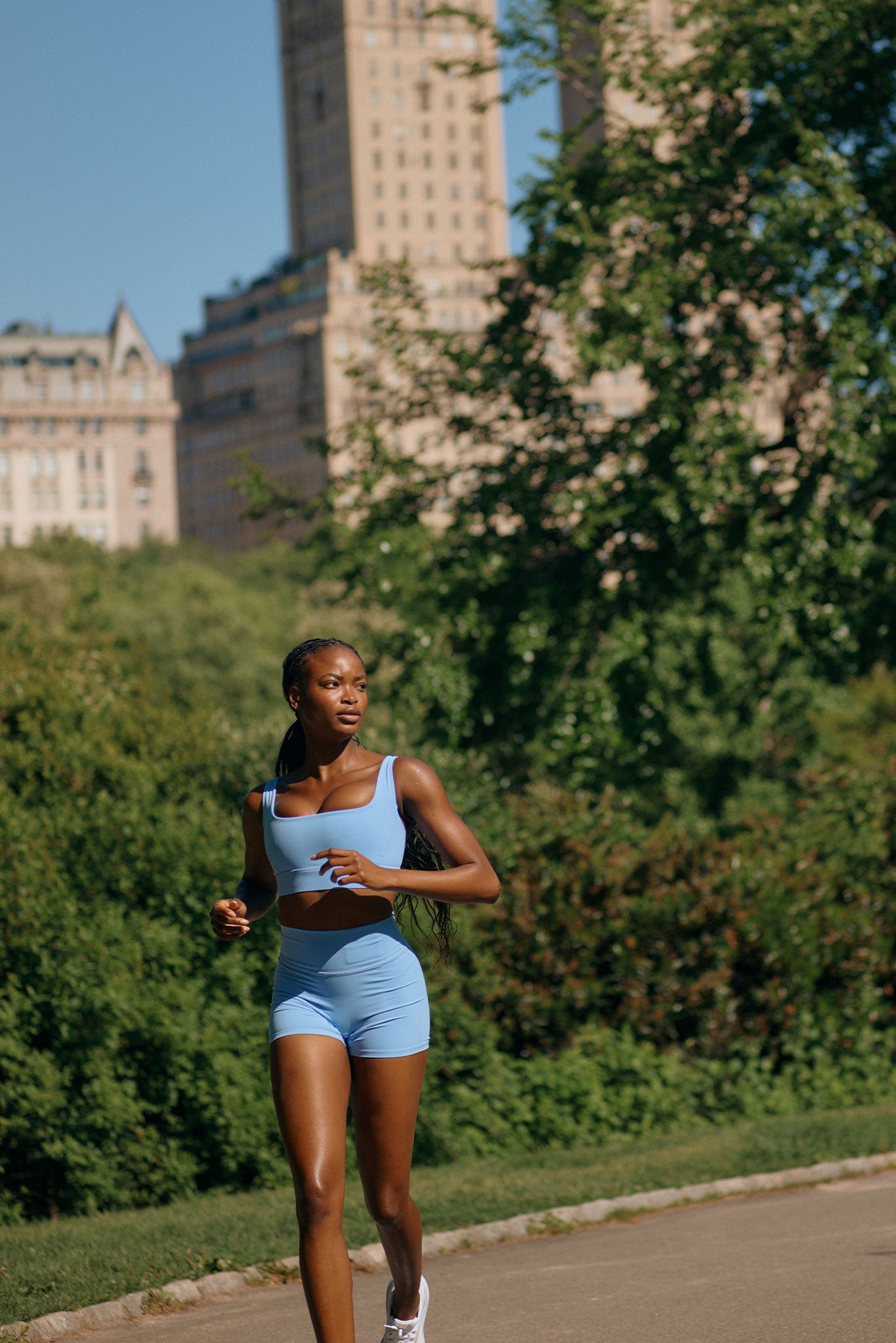 Woman jogging on a park path wearing light blue sports top and shorts, trees and city buildings in background, sunny day