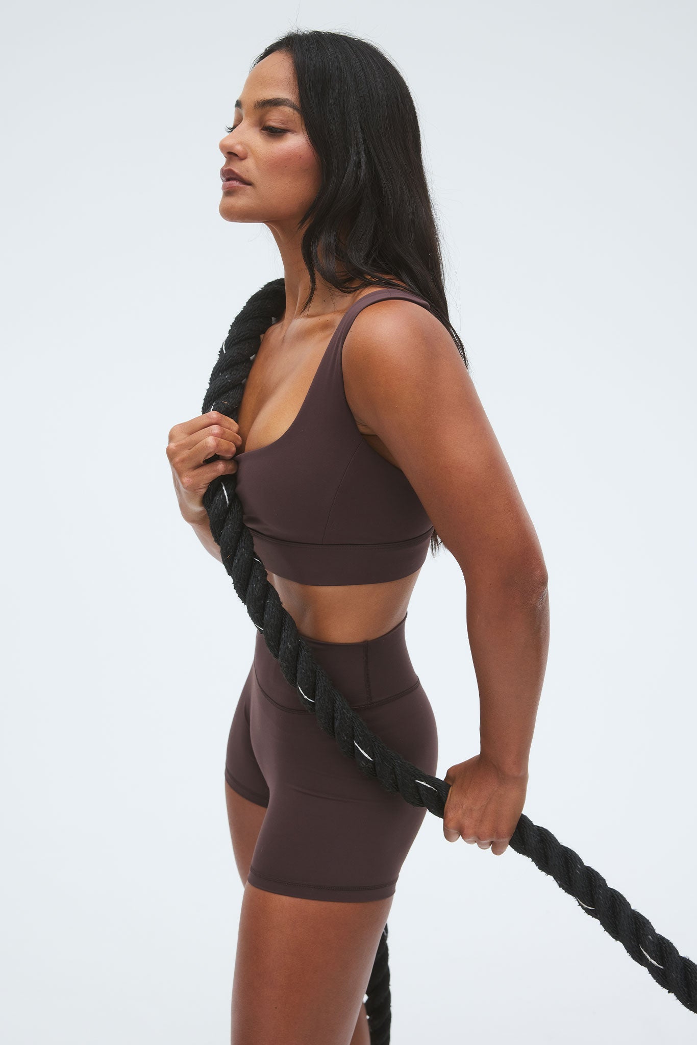 Woman in brown sports bra and shorts holding a thick black battle rope, side profile on a light background.
