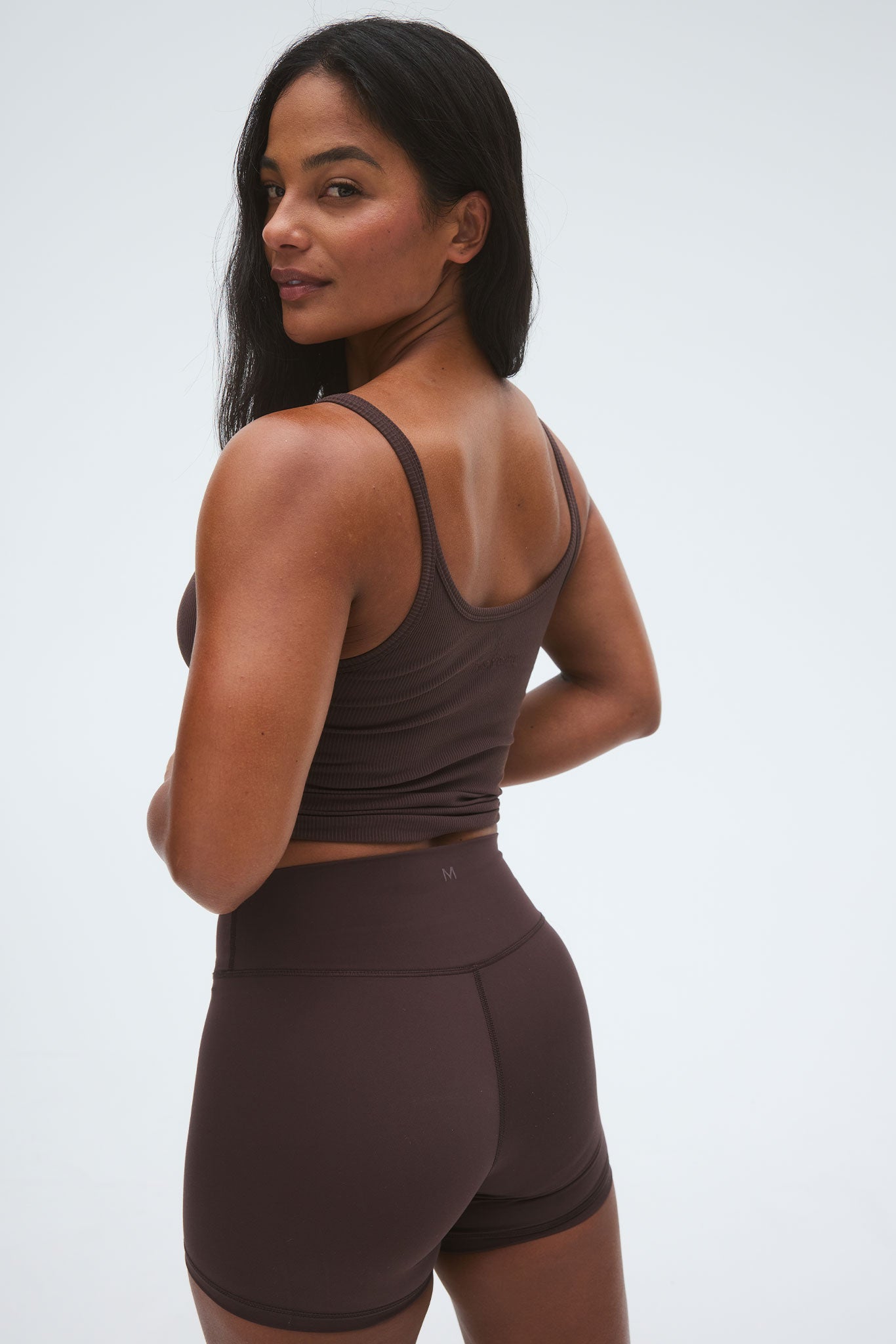 Woman wearing a brown ribbed crop tank and high-waist brown bike shorts, looking over her left shoulder.