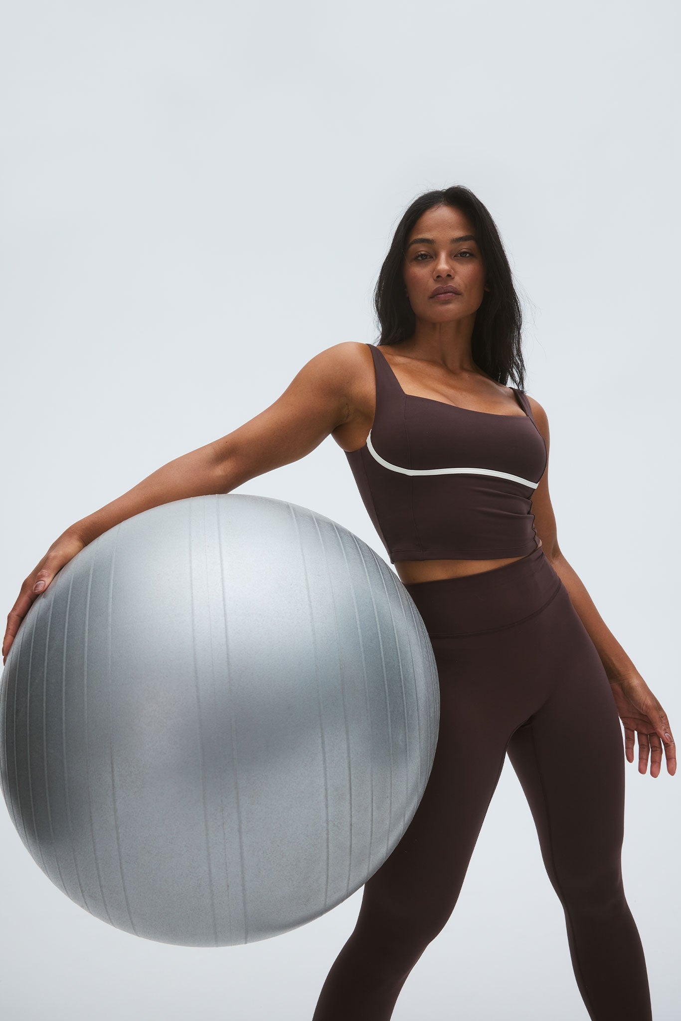 Woman in brown workout set leaning on a large silver exercise ball against a light gray background