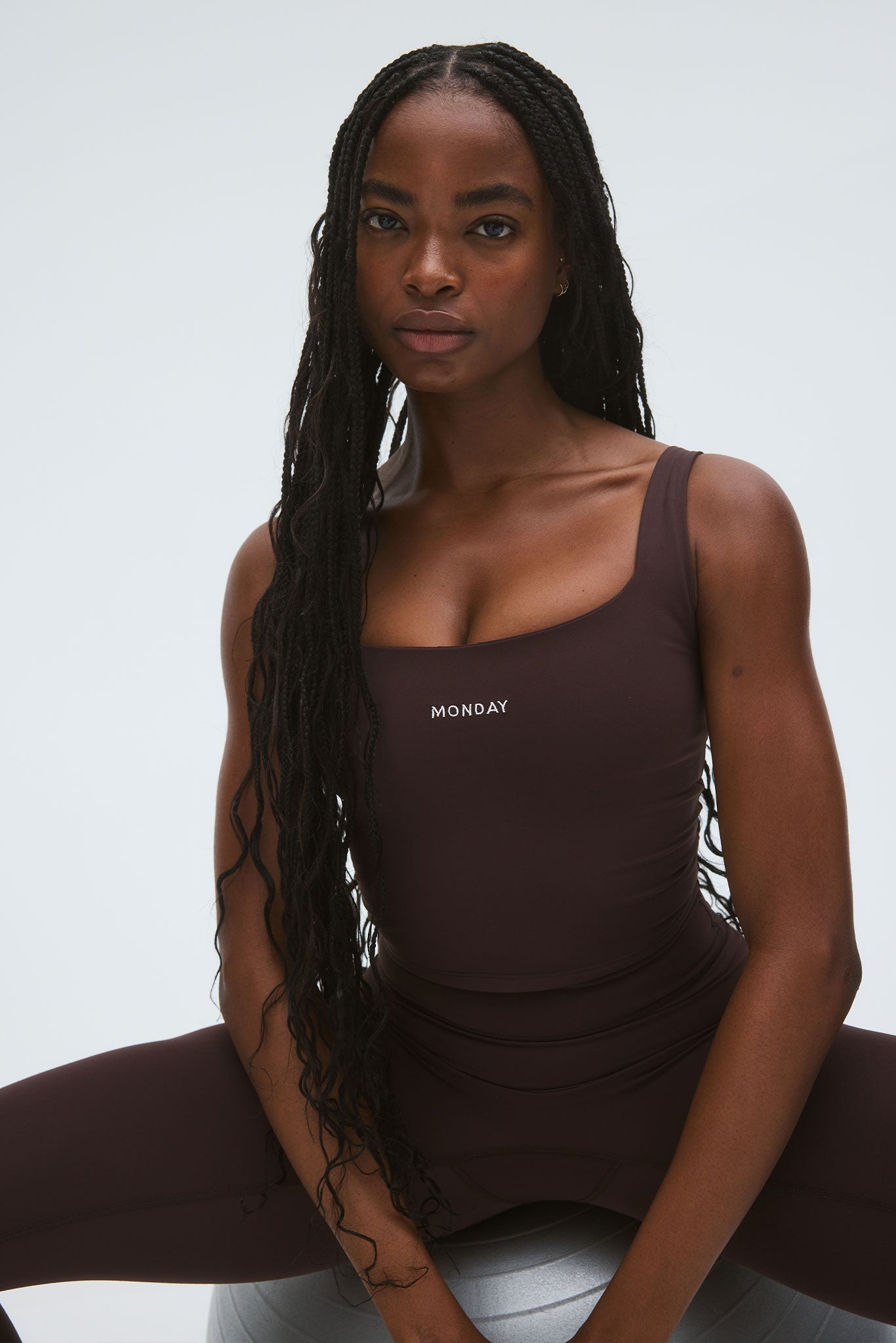 Black woman seated on exercise ball wearing brown tank top labeled MONDAY with long braided hair.