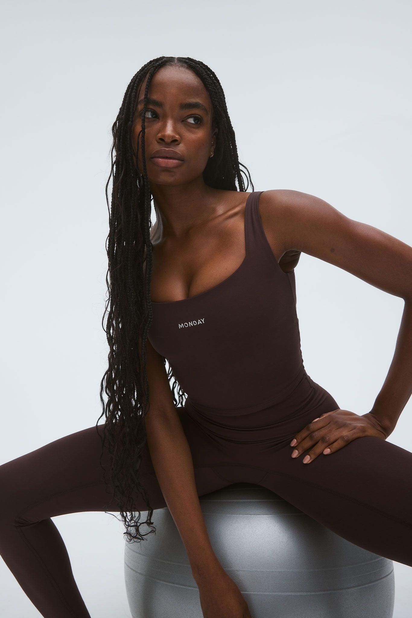 Woman with long braids in dark brown activewear bodysuit seated on a silver exercise ball, MONDAY printed on chest.