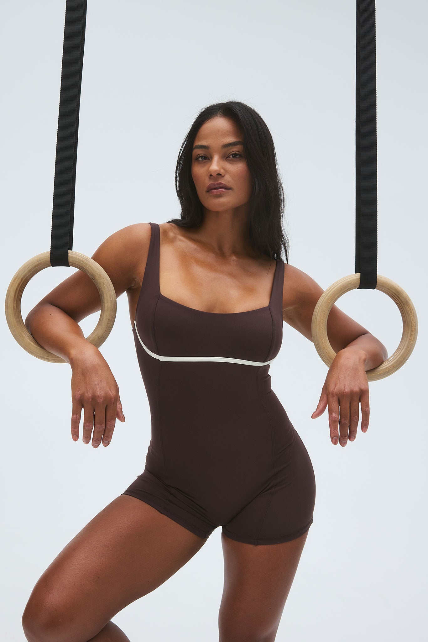 Model in brown athletic one-piece romper, arms draped over wooden gymnastic rings suspended from black straps.