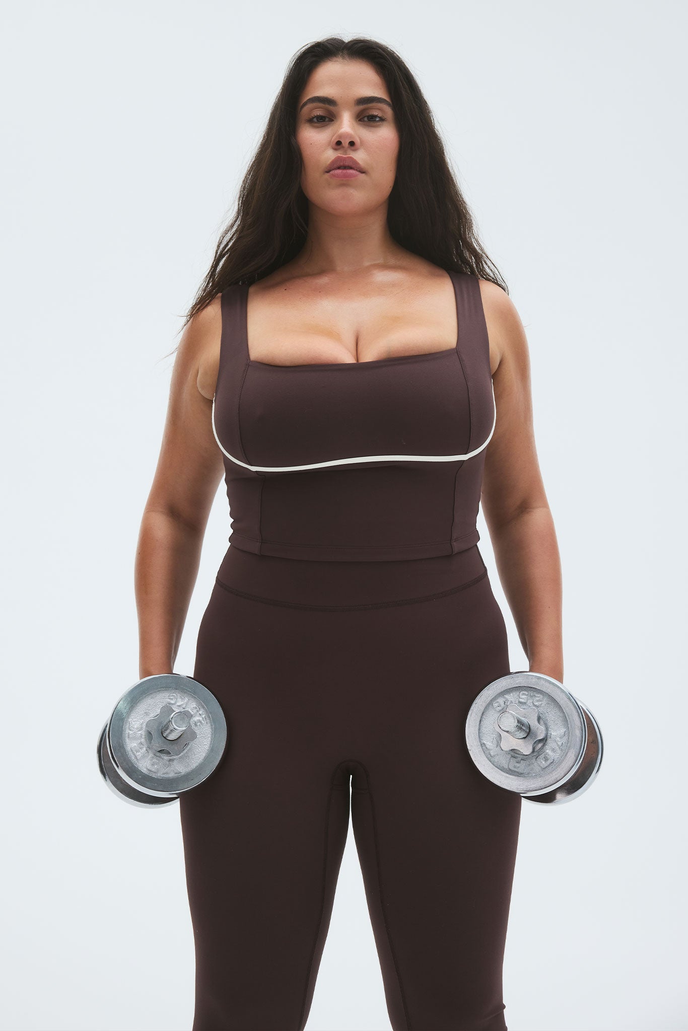 Person wearing brown workout crop top and leggings holding metal dumbbells at her sides in a frontal studio portrait.