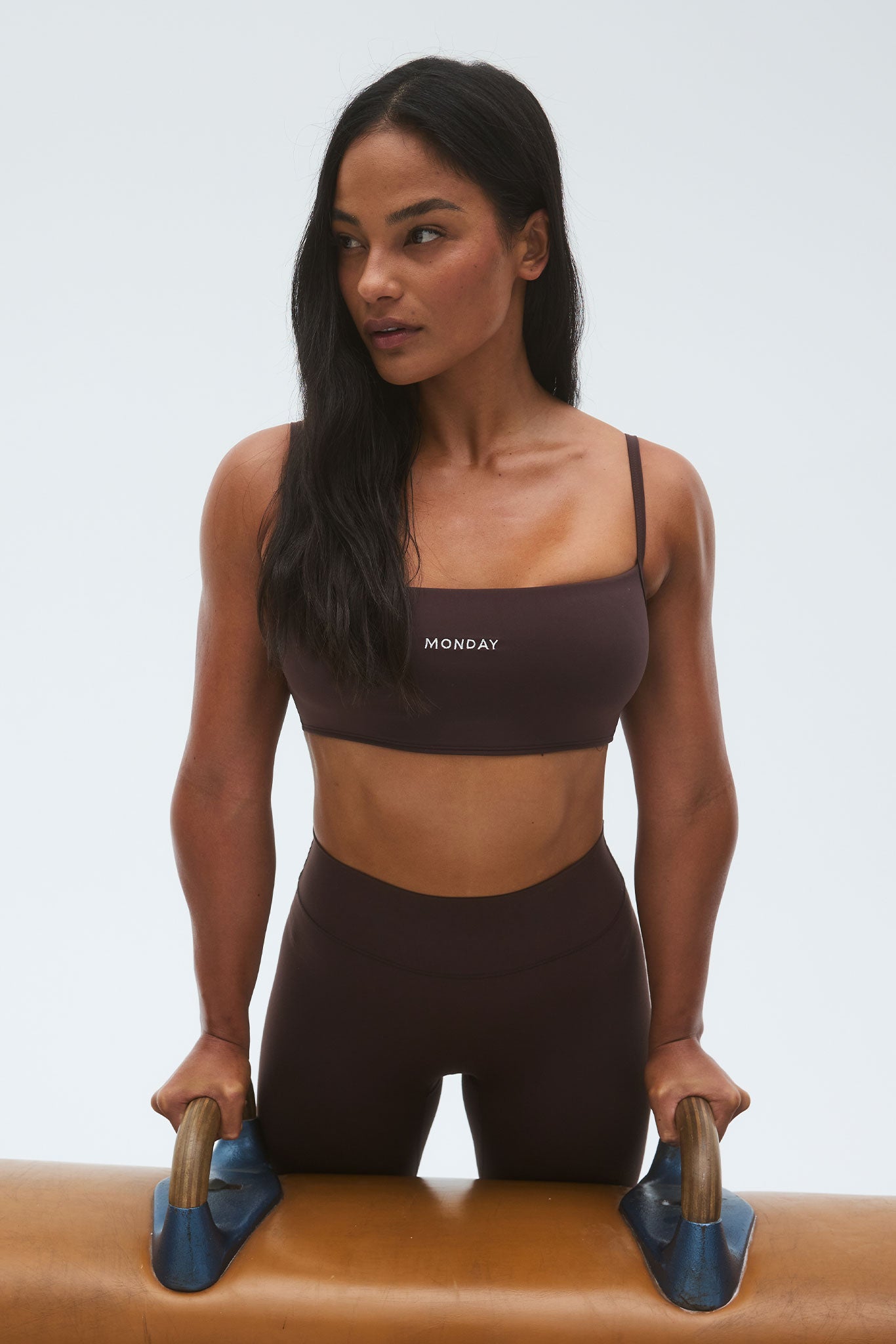 Athletic woman in dark brown sports bra and leggings, hands on pommel horse handles; 'MONDAY' printed on the bra.