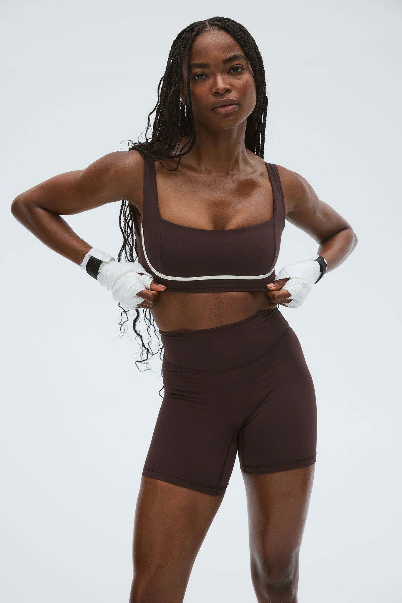Fit woman in dark brown sports bra and high-waist biker shorts, adjusting top with white hand wraps on gray background.
