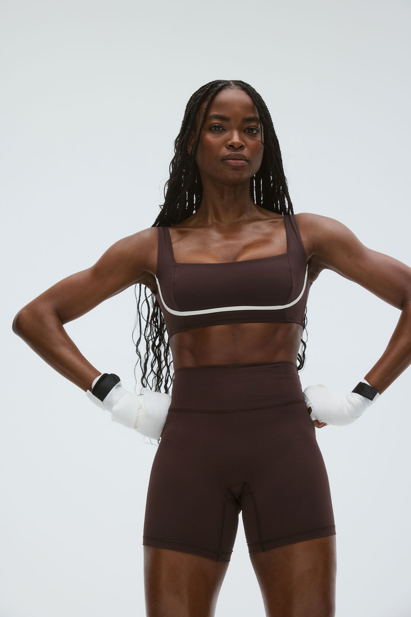 Female athlete standing with hands on hips wearing brown sports bra and high-waisted shorts and white hand wraps.