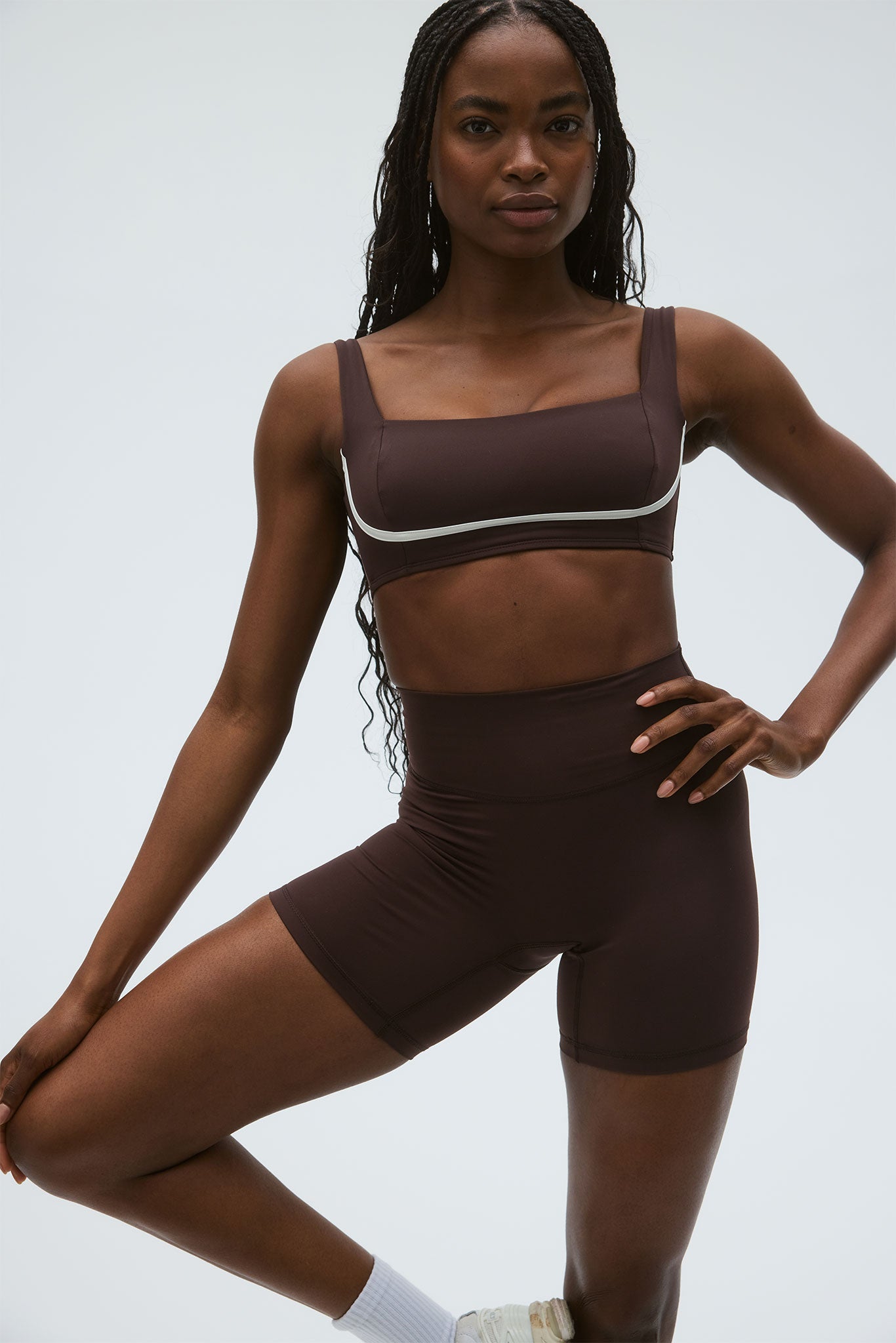 Model wearing brown square-neck sports bra and high-waist biker shorts, hand on hip and one knee lifted