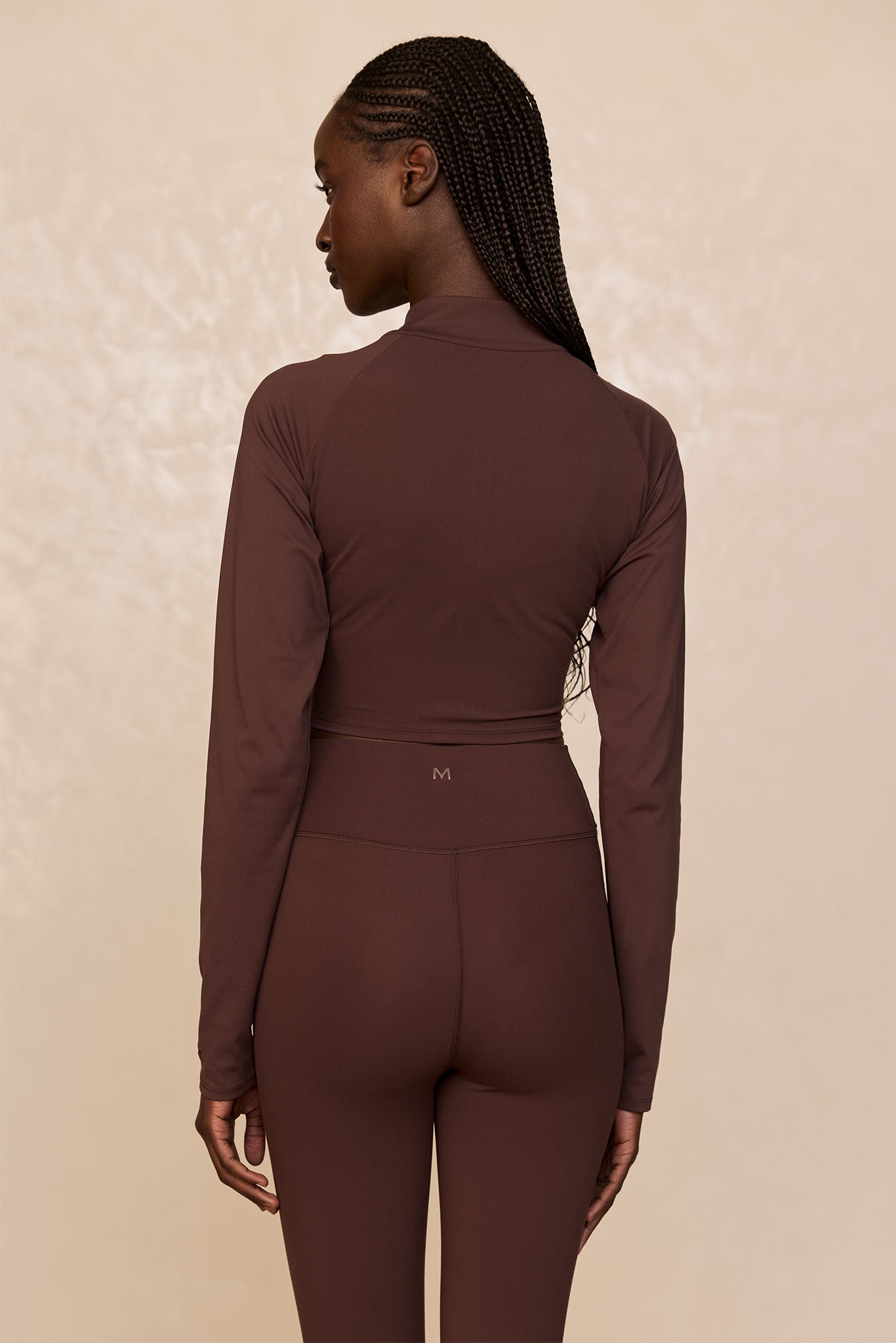 Rear view of woman wearing brown long-sleeve crop top and high-waist leggings; small M logo on waistband.