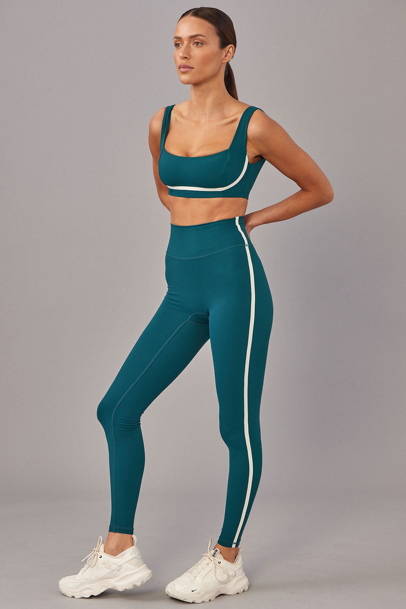 Model wearing teal sports bra and high-waist leggings with white side stripe, standing in white sneakers