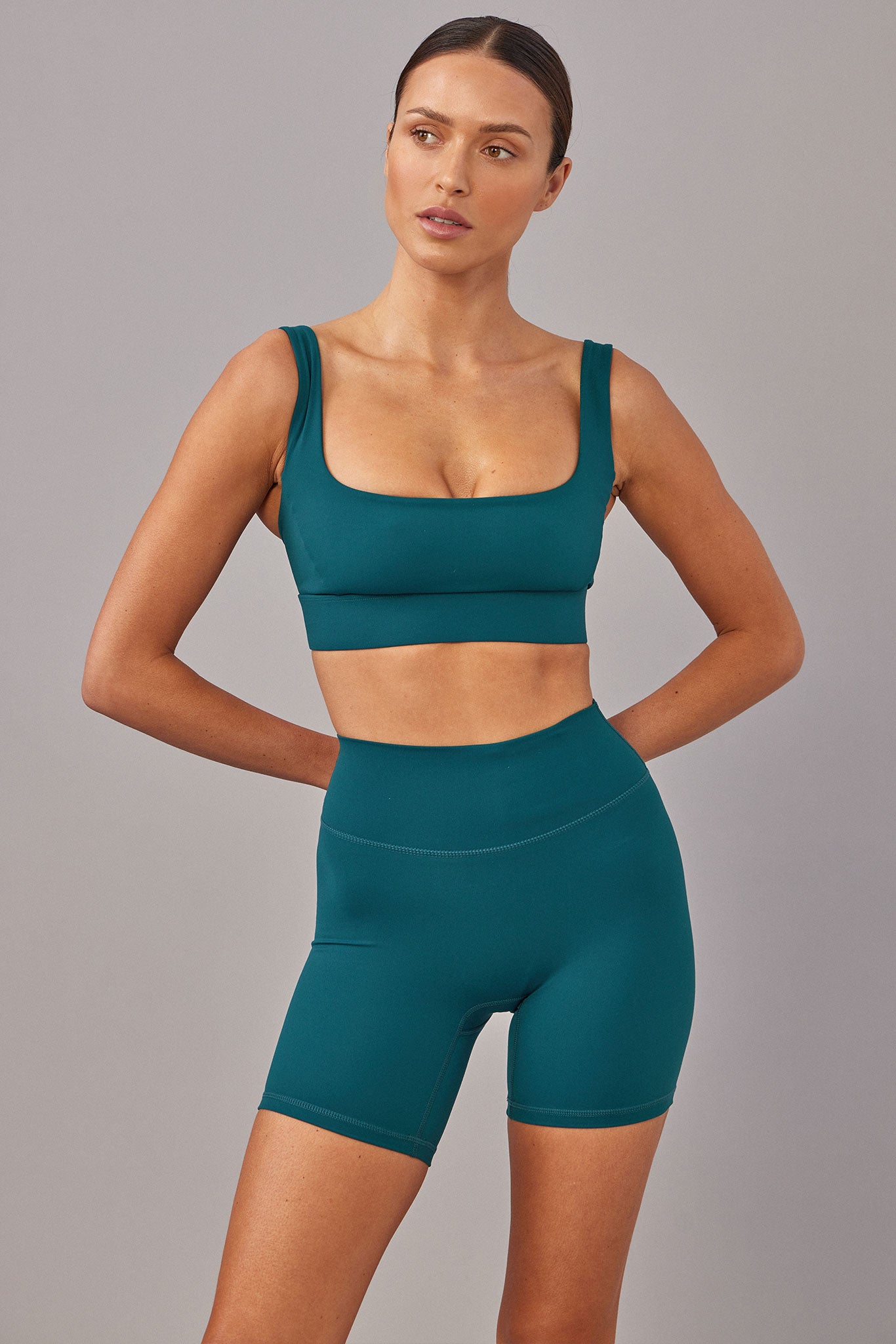 Model wearing teal square-neck sports bra and high-waist biker shorts, hands behind hips, gray background.