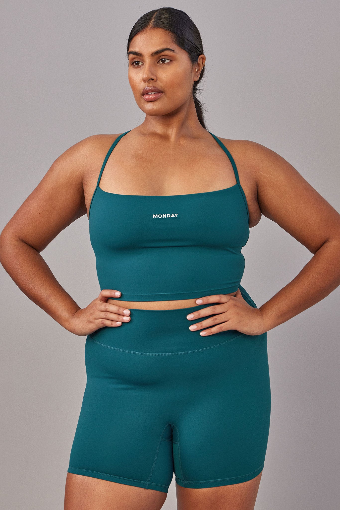 Plus-size model wearing teal crop sports bra and high-waist bike shorts; 'MONDAY' logo centered on the bra.