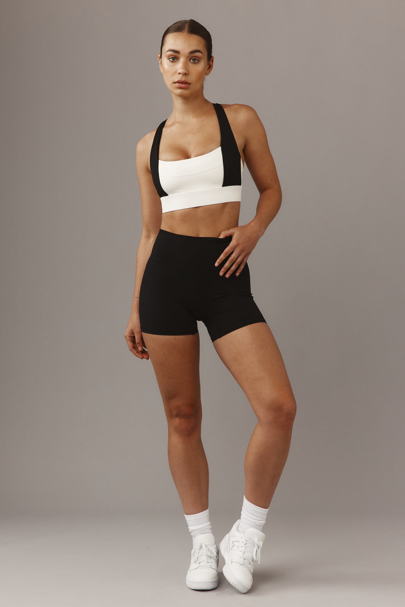 Woman standing in white sports bra with black straps, black high‑waisted shorts and white sneakers on gray background.