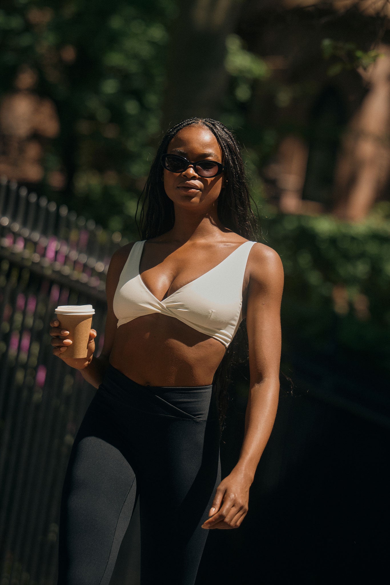 Woman in sunglasses and white crop top with black leggings, walking outdoors holding a takeaway coffee cup in sunlight.