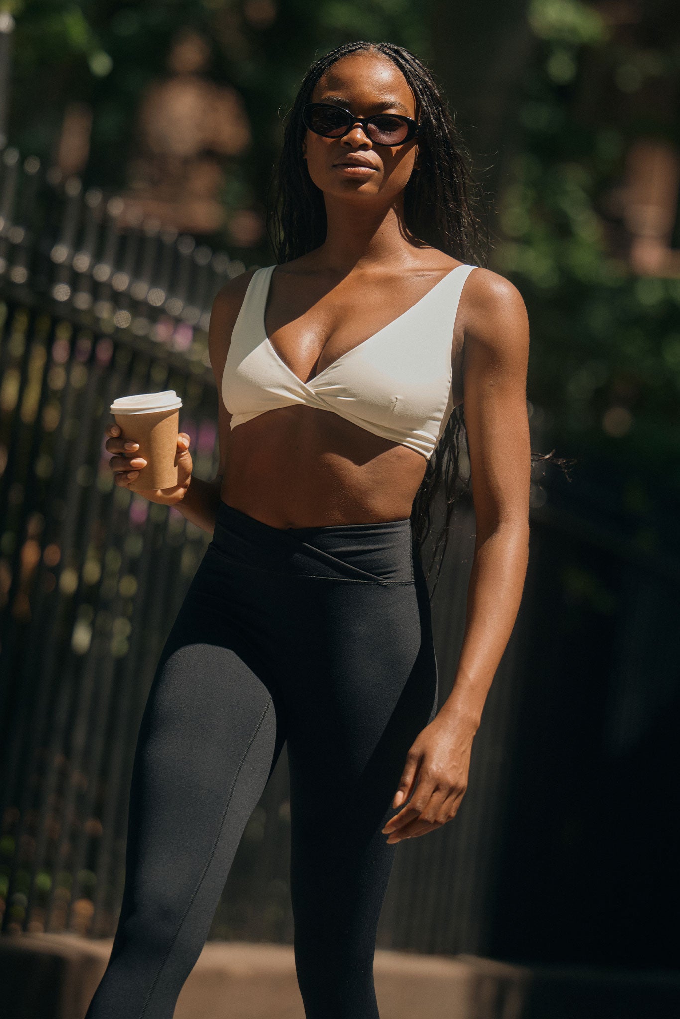 Woman walking outdoors in sunglasses, cream twist-front crop top and high-waist black leggings, holding a paper coffee cup.
