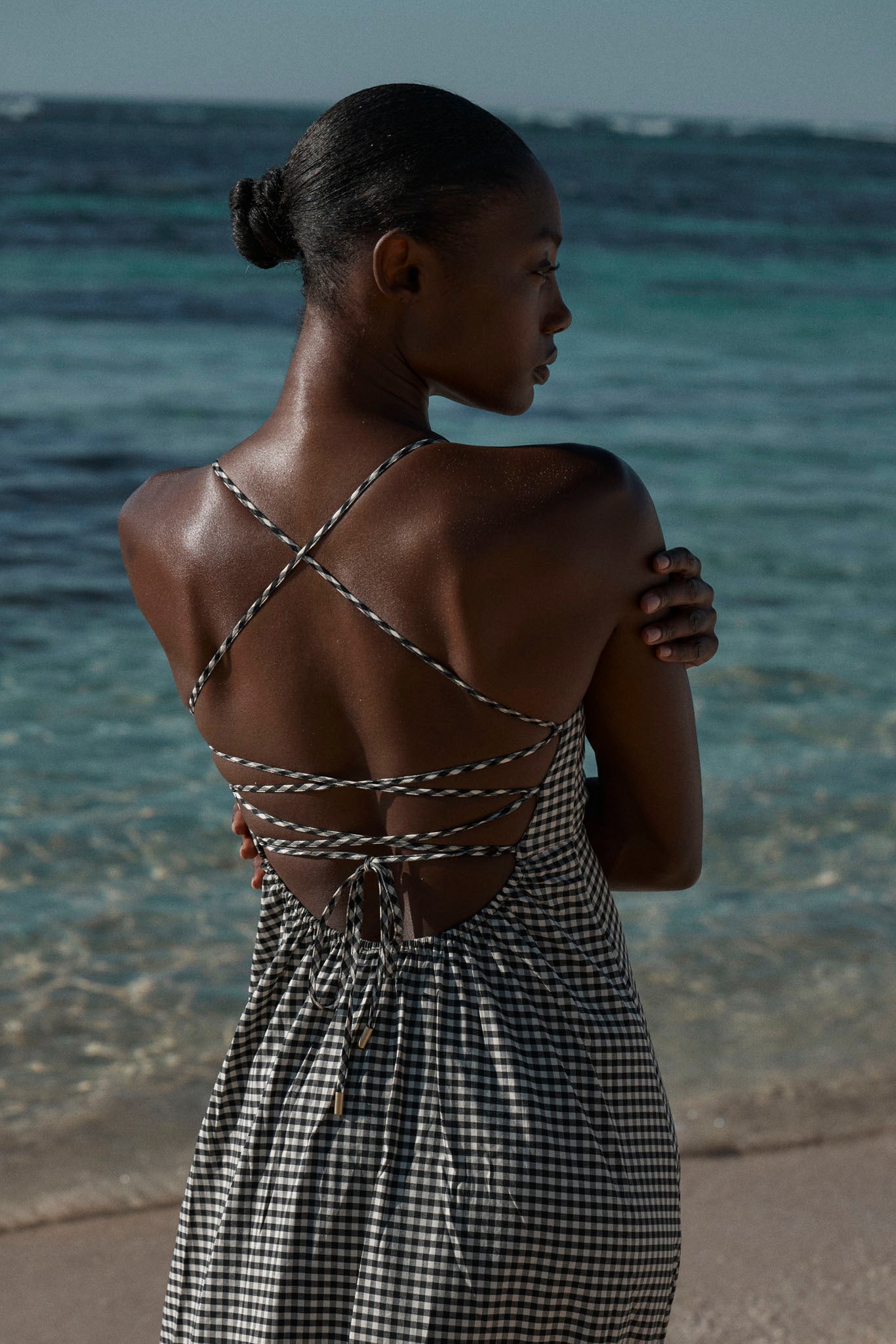 Rear view of a woman in a black-and-white gingham sundress with crisscross straps standing at the shoreline