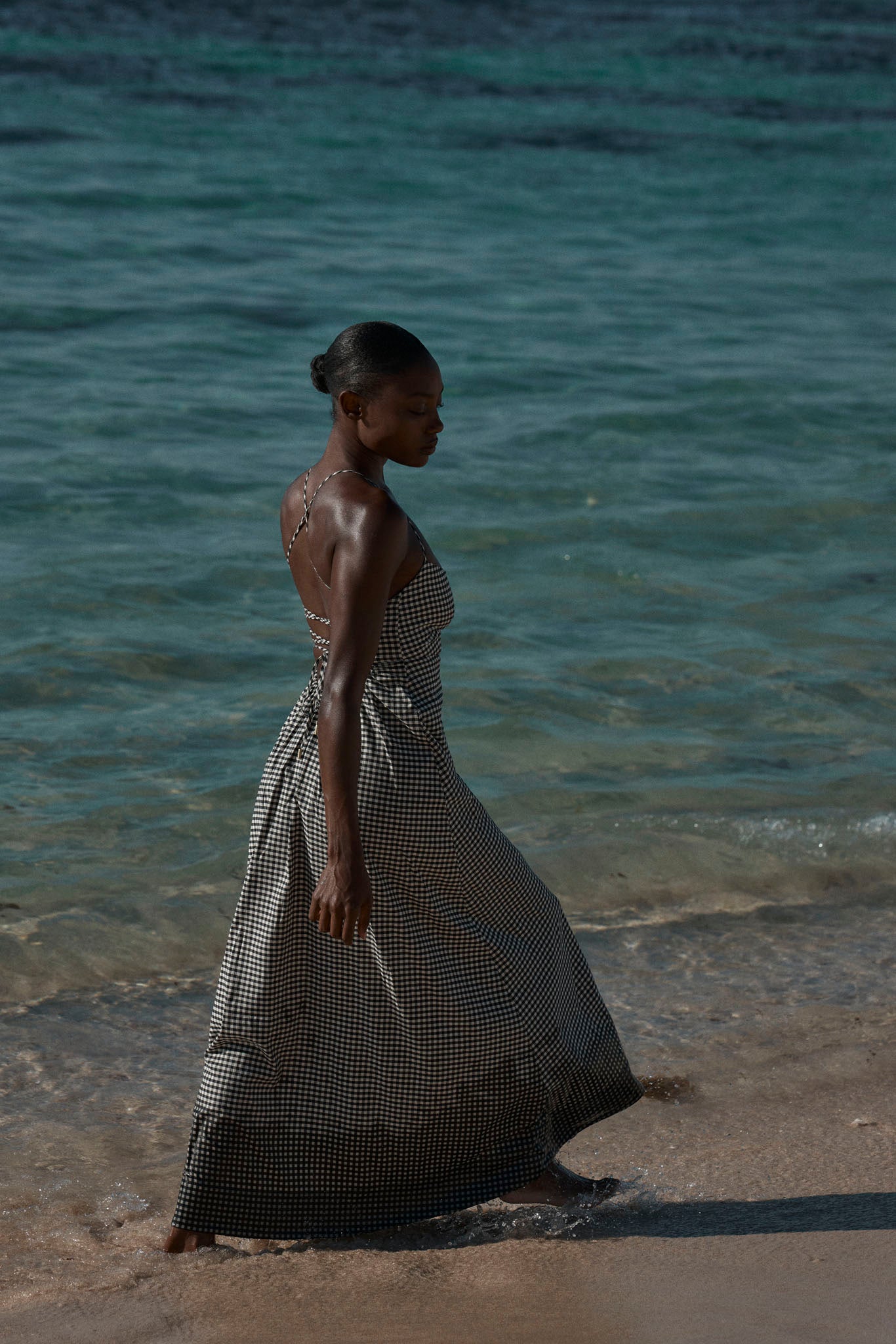 Woman in a black-and-white gingham maxi dress walking along a sandy shore with turquoise sea