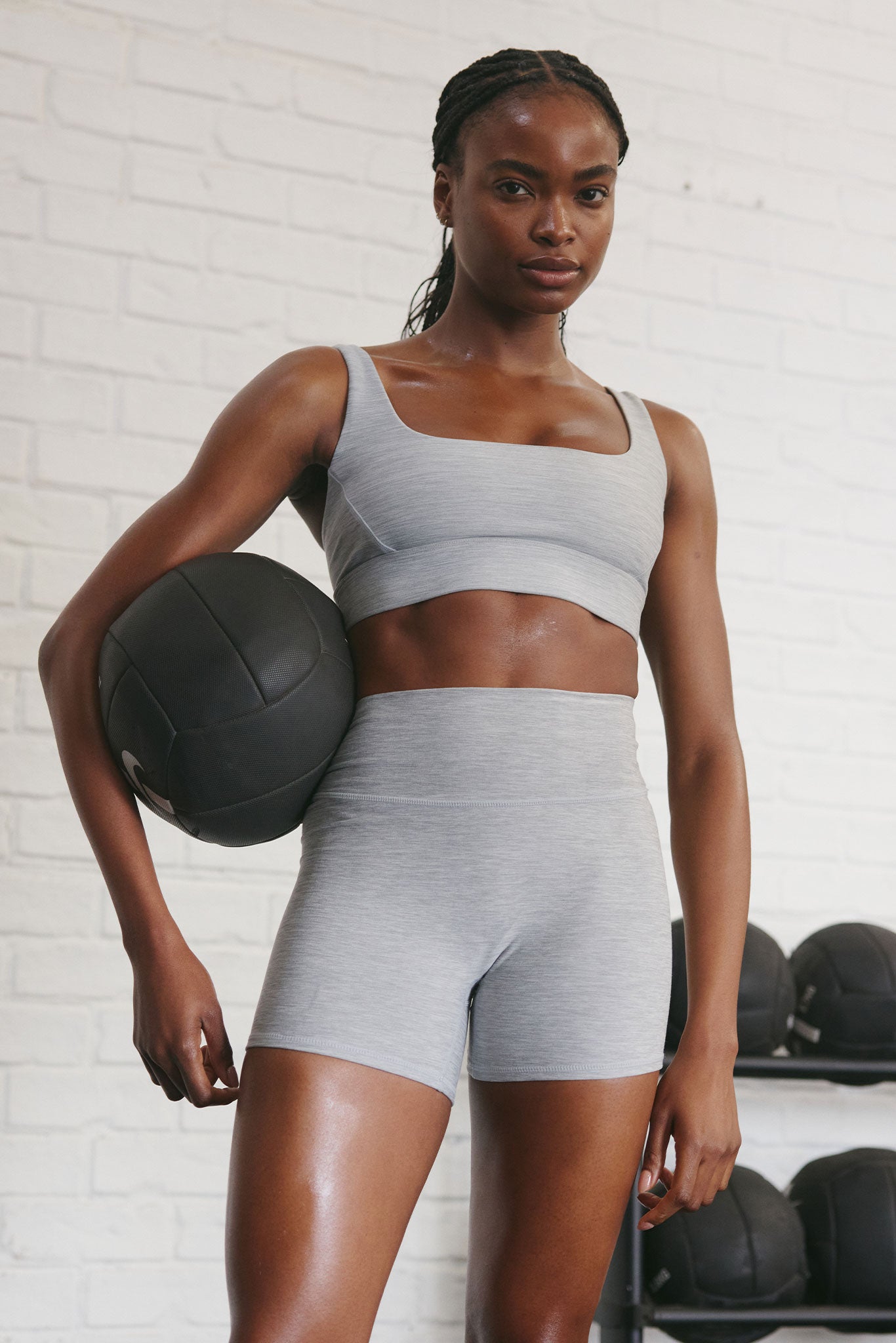 Athletic woman in gray sports bra and high-waist shorts holding a black medicine ball by a white brick gym wall.