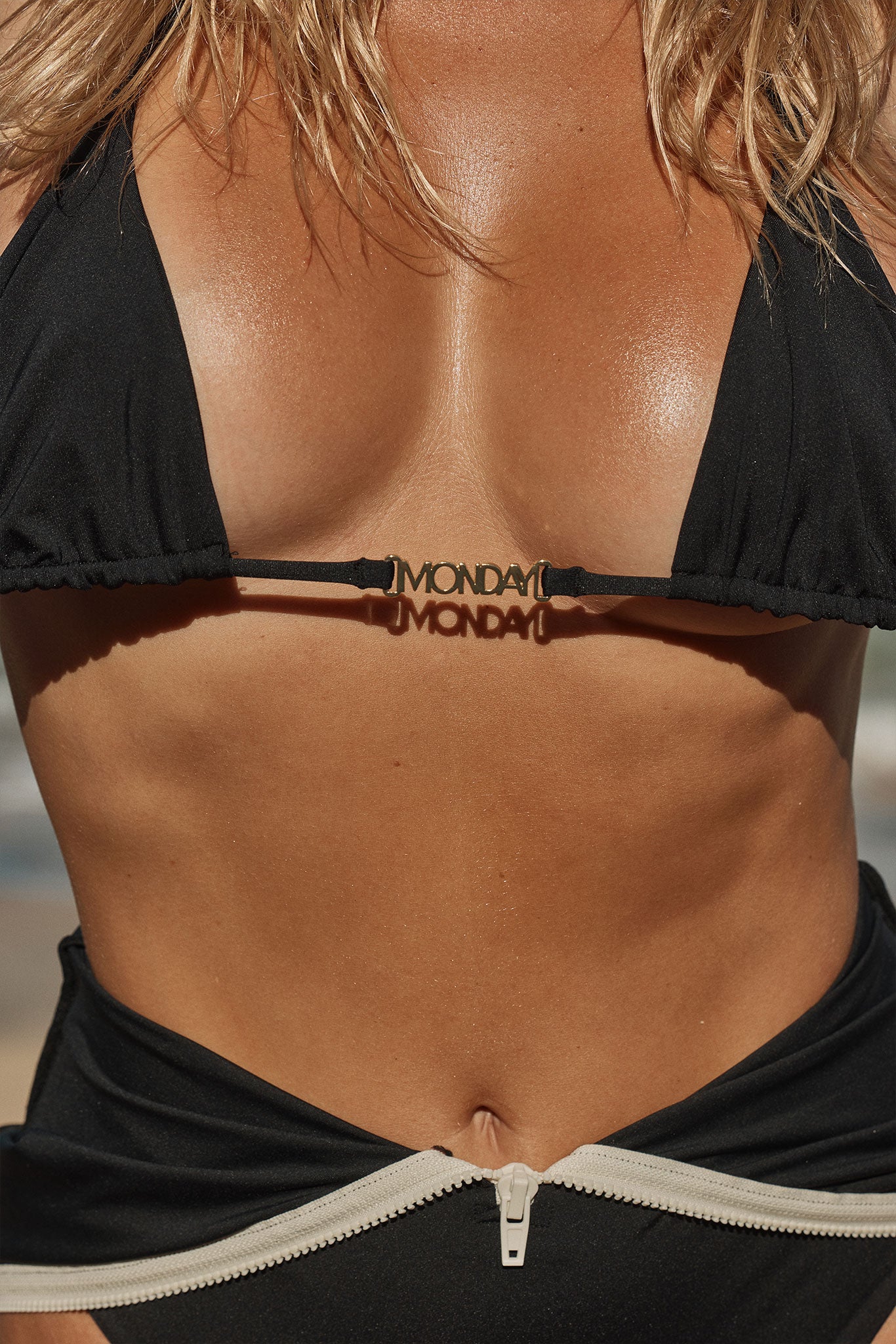 Tanned female torso in a black bikini with a gold "MONDAY" nameplate at the center strap.